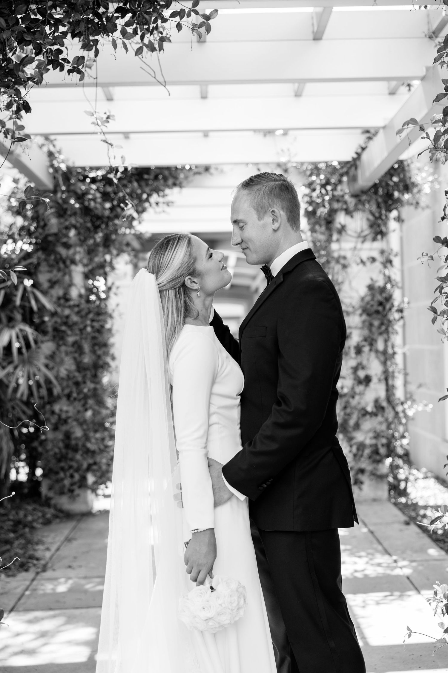 Bride and groom share intimate moment under white pergola with lush greenery in black and white portrait.