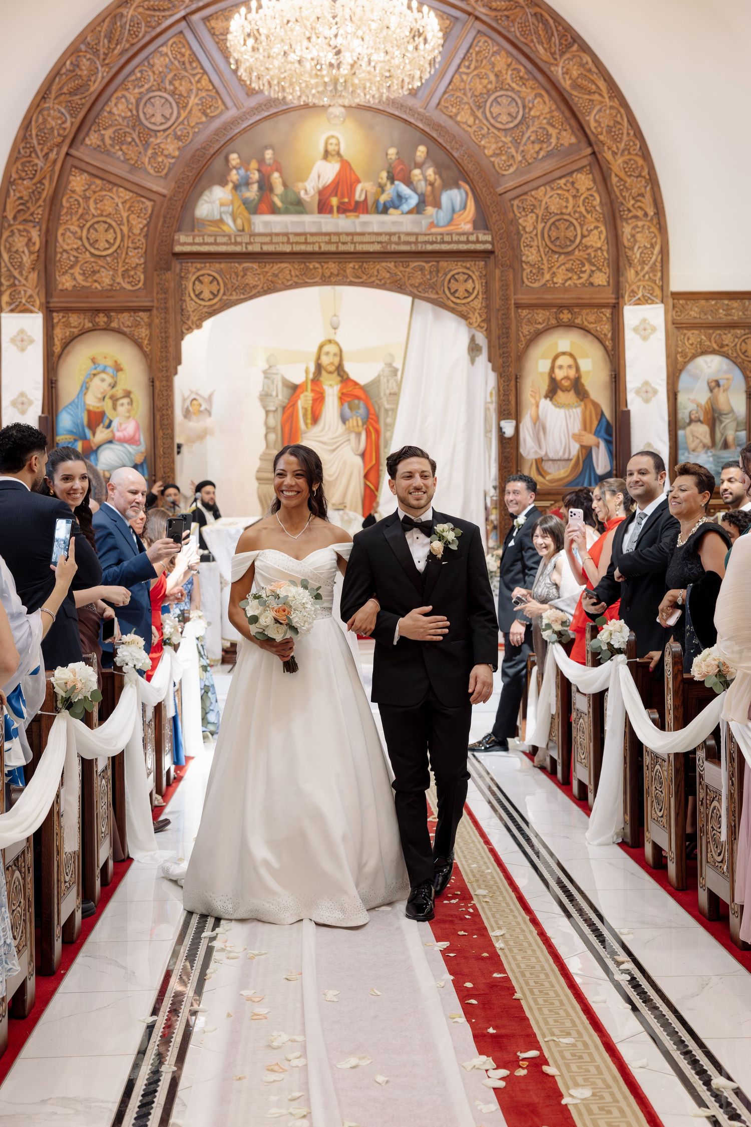 Newlywed couple walks down ornate church aisle after ceremony beneath religious murals and chandelier.