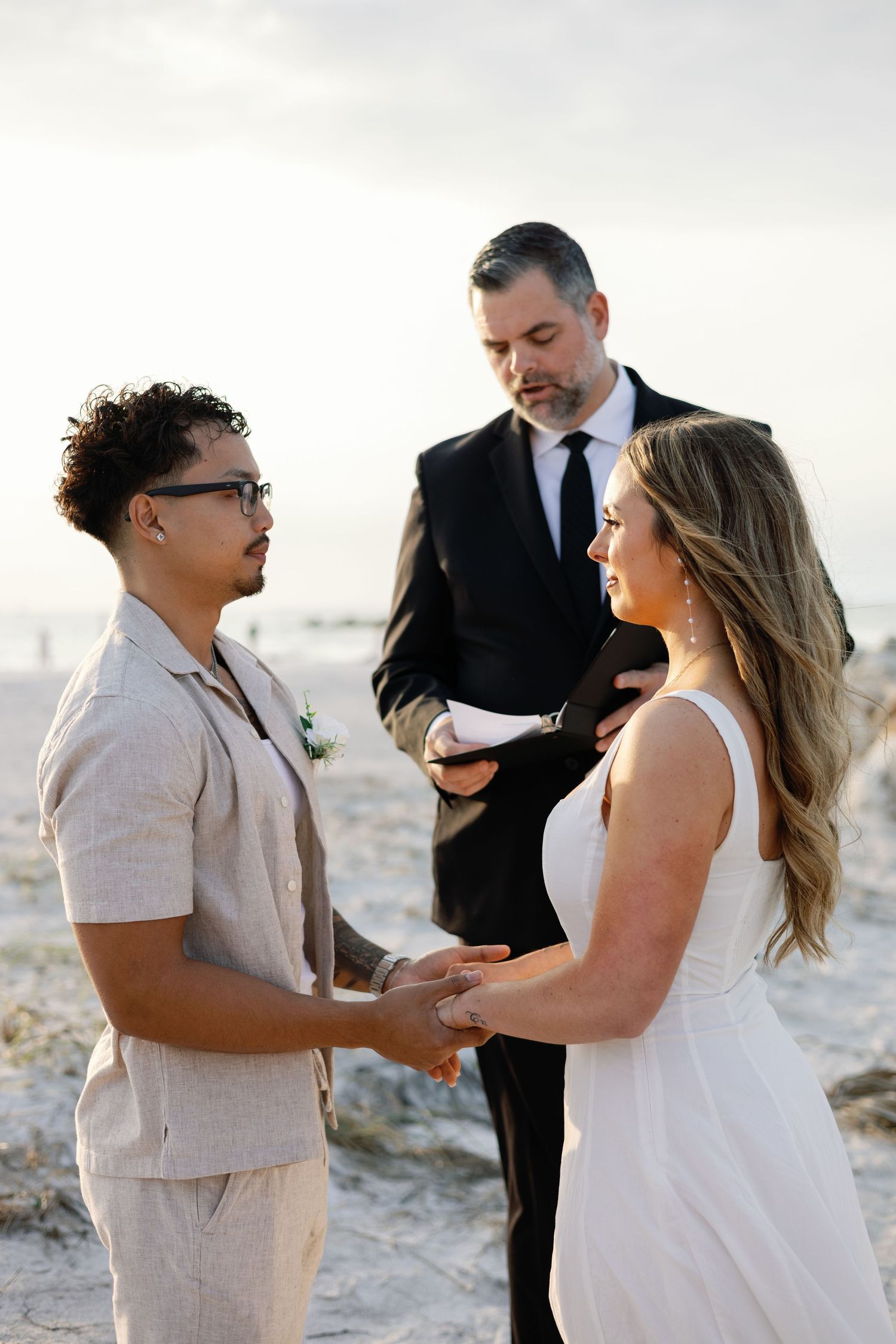 Couple exchanges vows holding hands before officiant during sunset beach wedding.