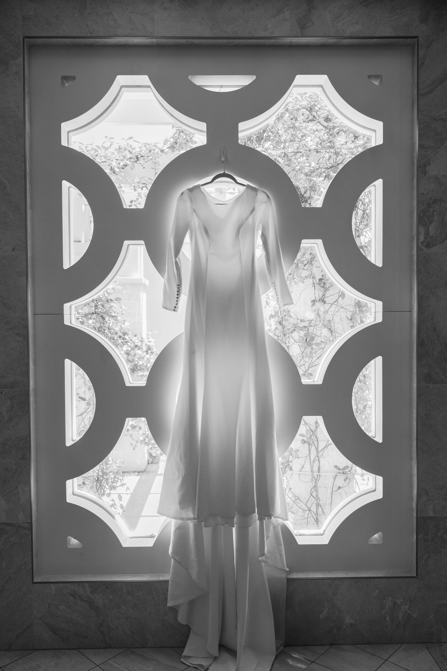 Black and white photo of elegant long-sleeve wedding gown hanging in ornate decorative window.