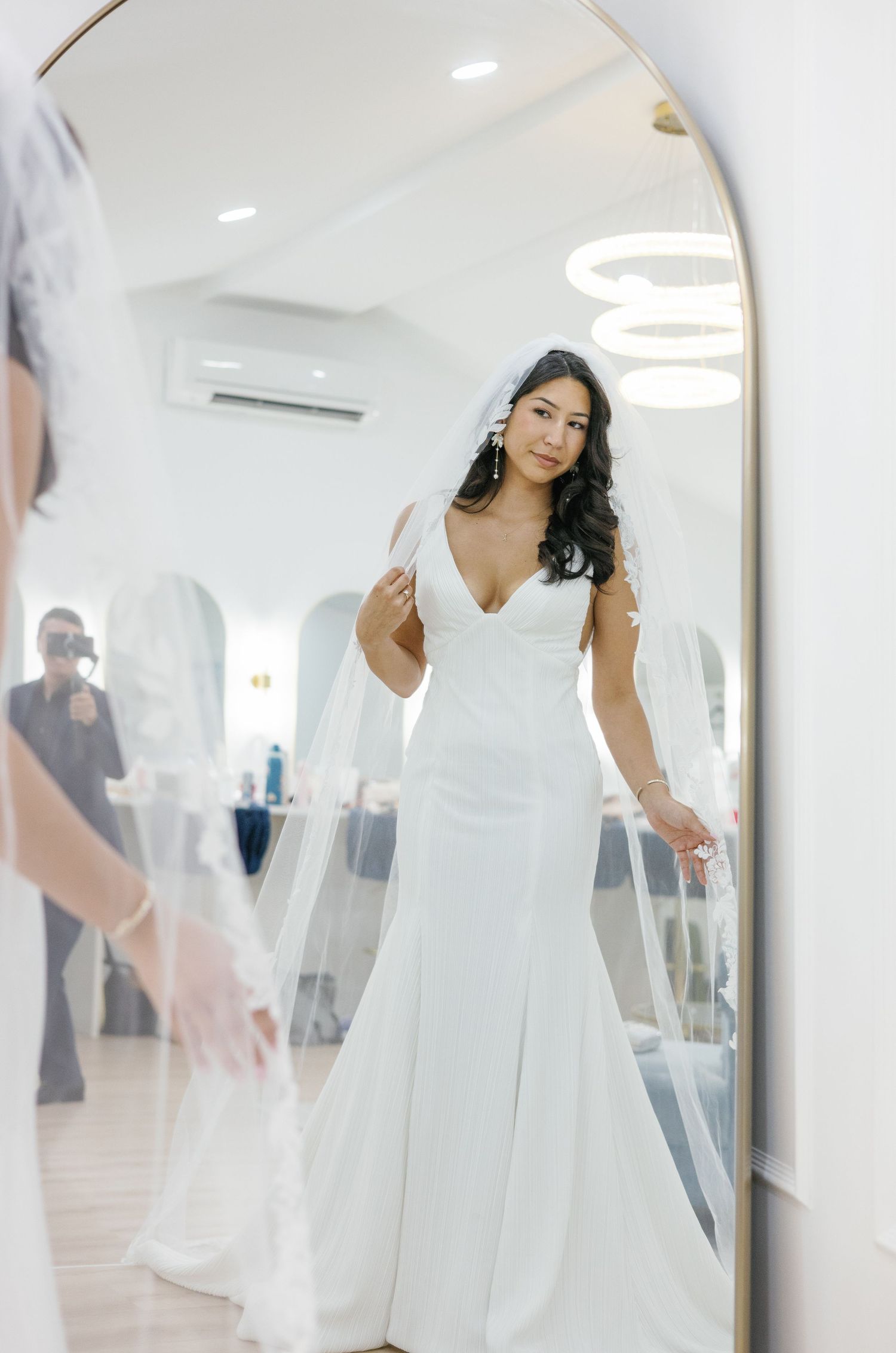 Bride in sleek white gown and veil poses in arched mirror reflection in bridal suite.