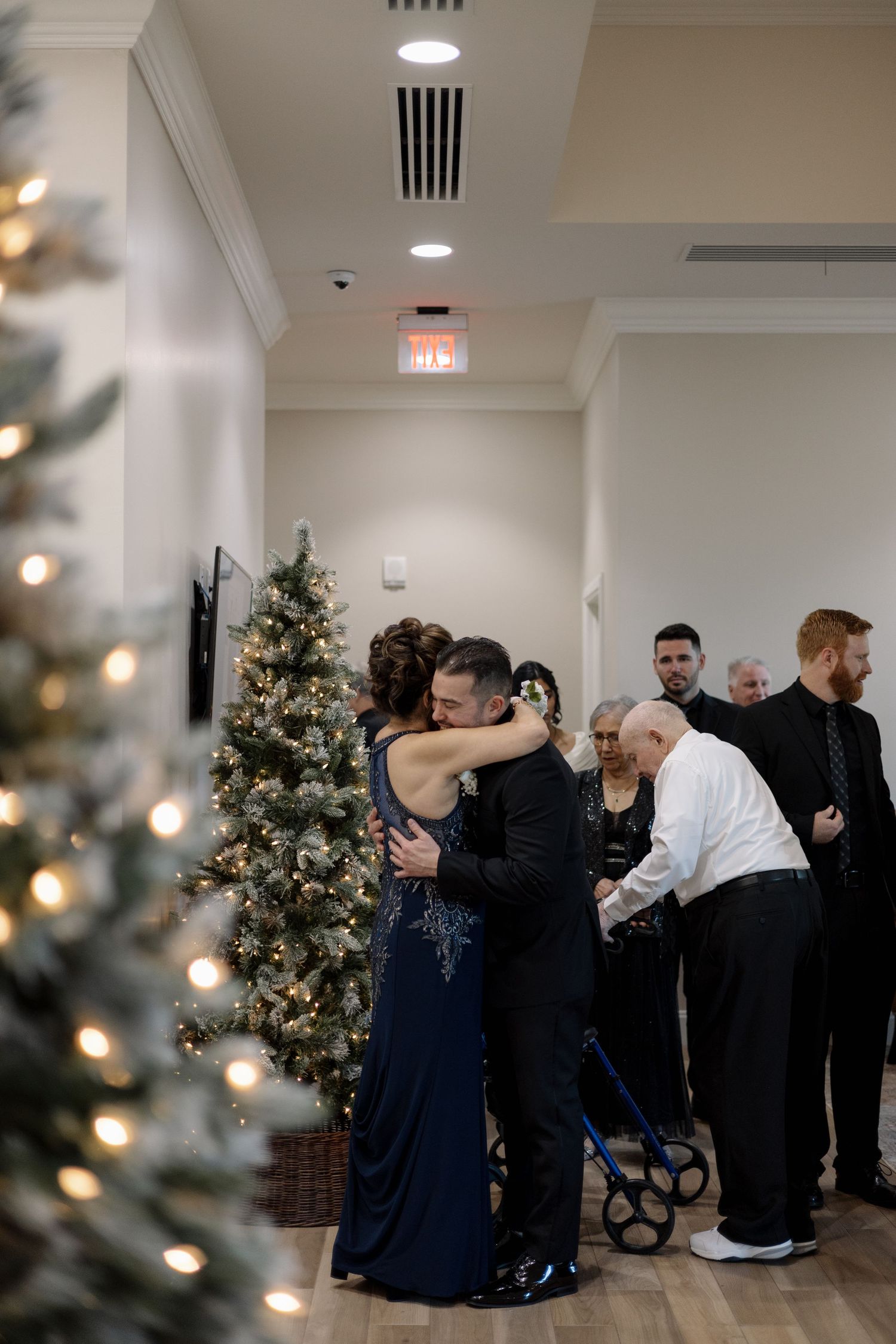 Wedding guests embrace near a decorated Christmas tree in a warmly lit venue hallway.