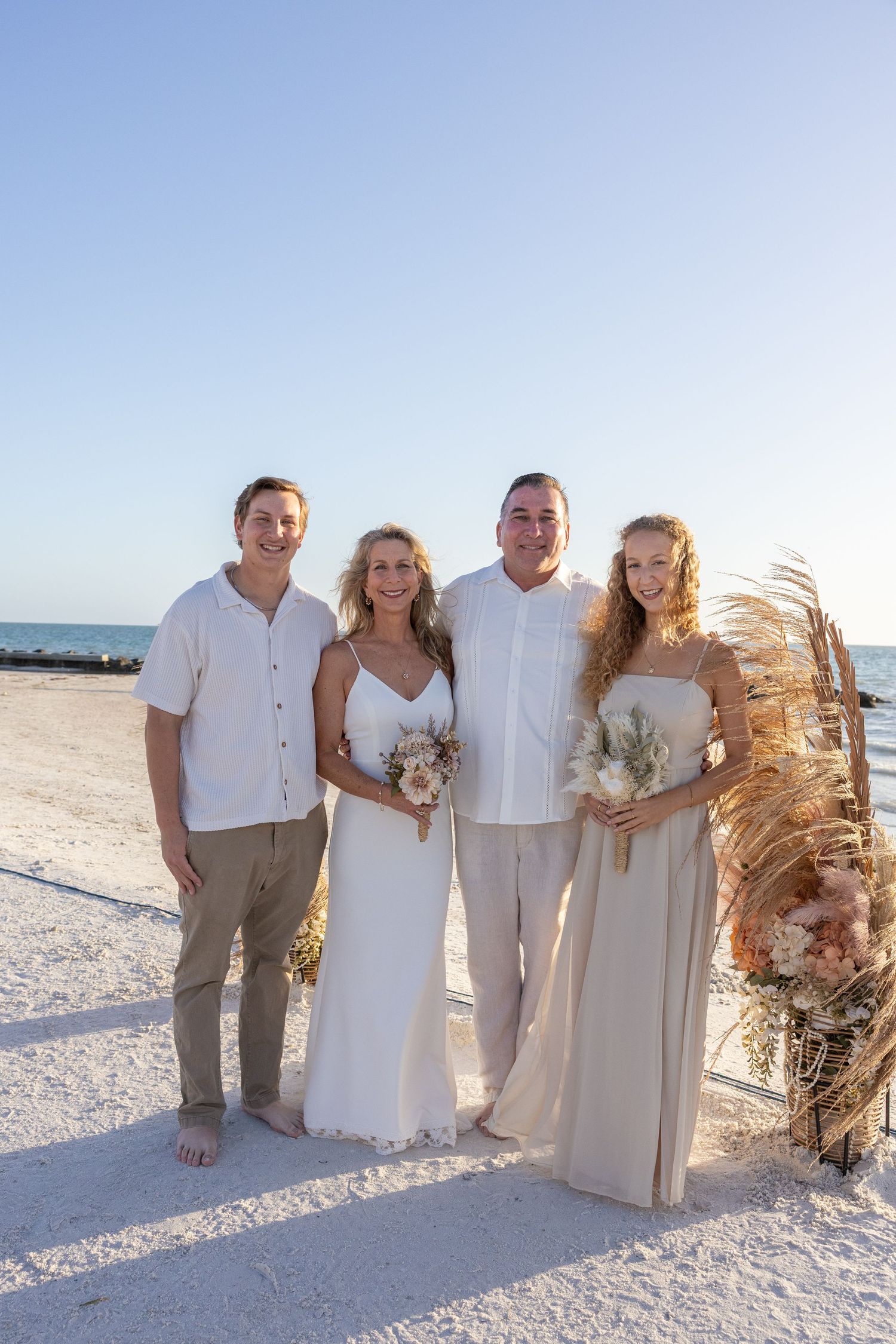 Newlywed couple poses with two guests on white sand beach after sunset wedding ceremony.