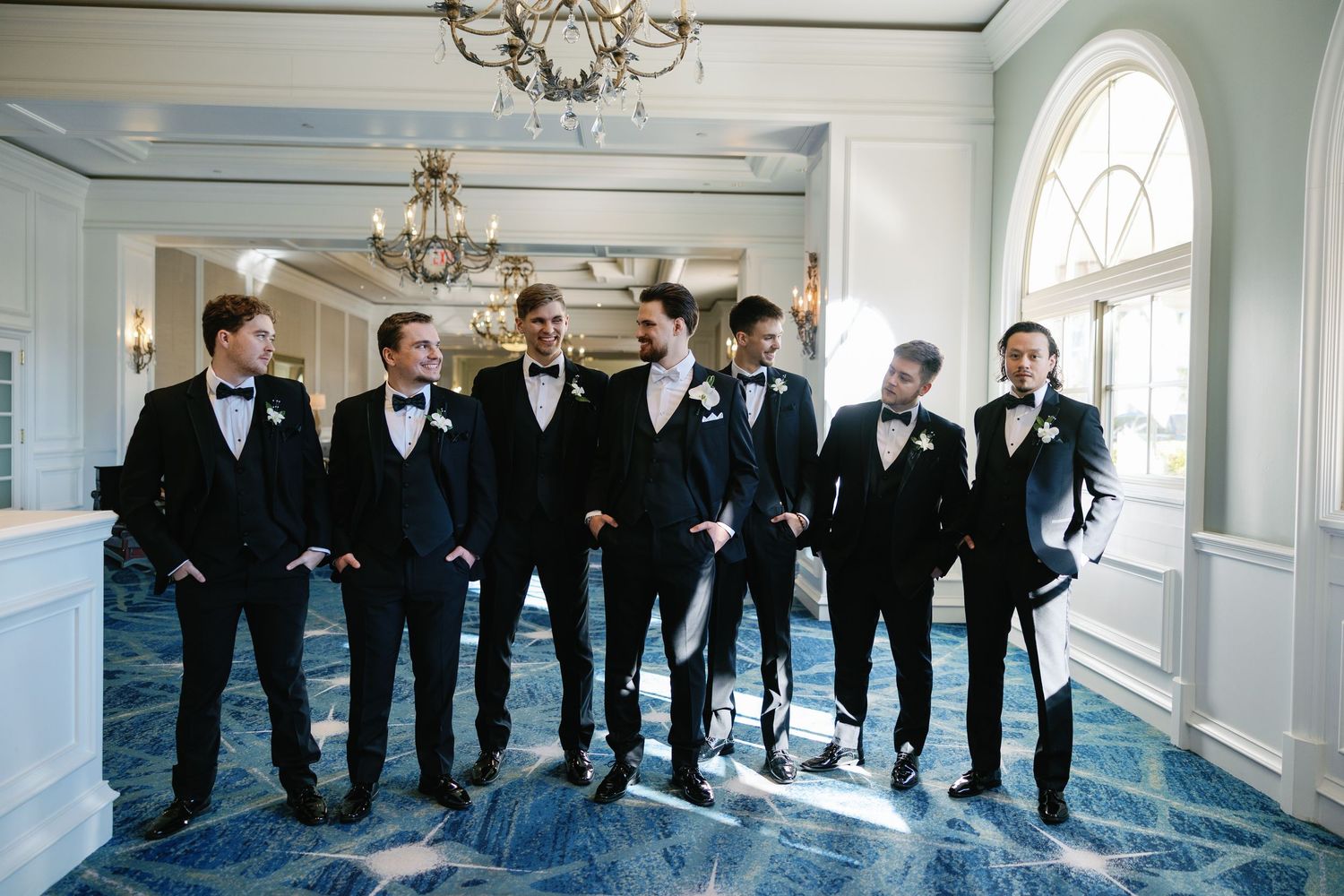 Groom and groomsmen in black tuxedos with bow ties posing in elegant wedding venue with blue floors.