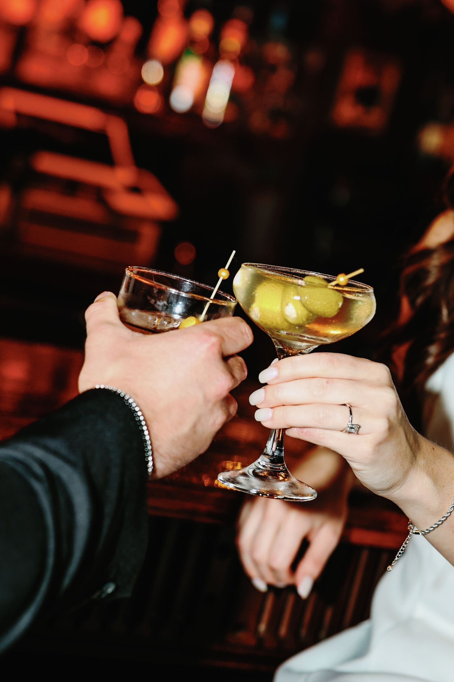 Engaged couple toasts with dirty martini cocktails garnished with olives in a dimly lit bar.