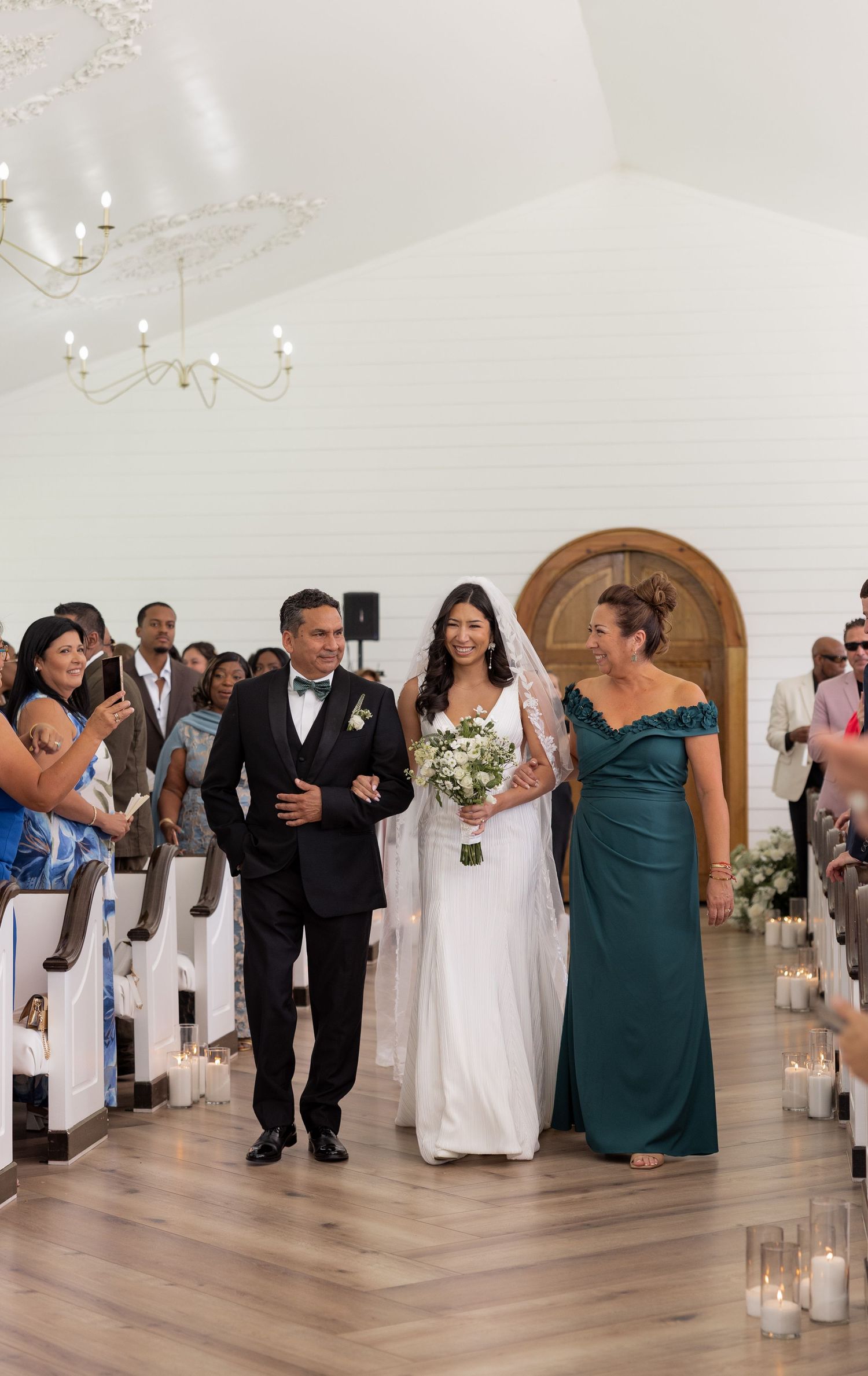Bride walks down aisle escorted by parents in elegant white chapel during wedding ceremony.