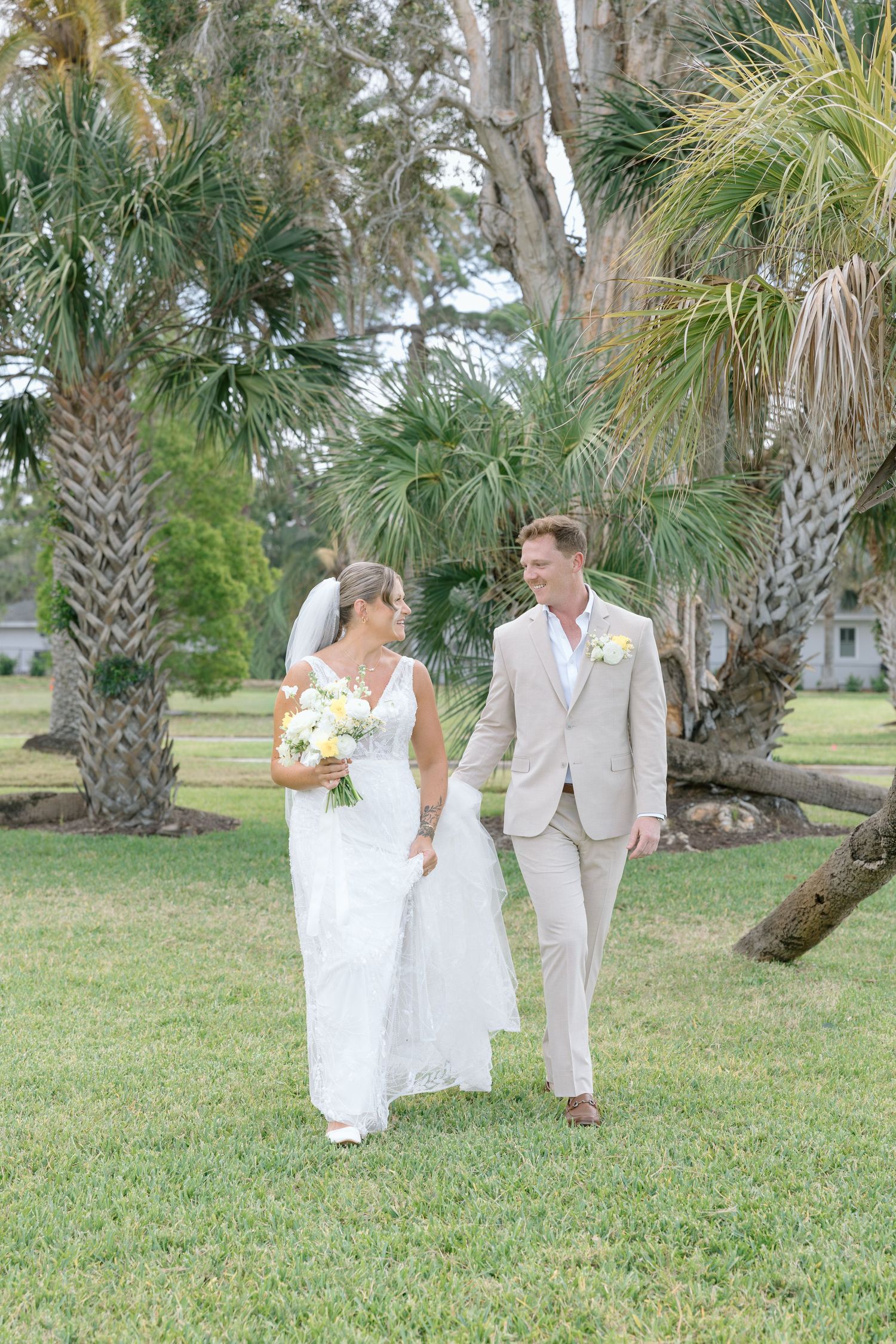 Bride in lace wedding gown and groom in tan suit walking hand in hand among tropical palm trees.