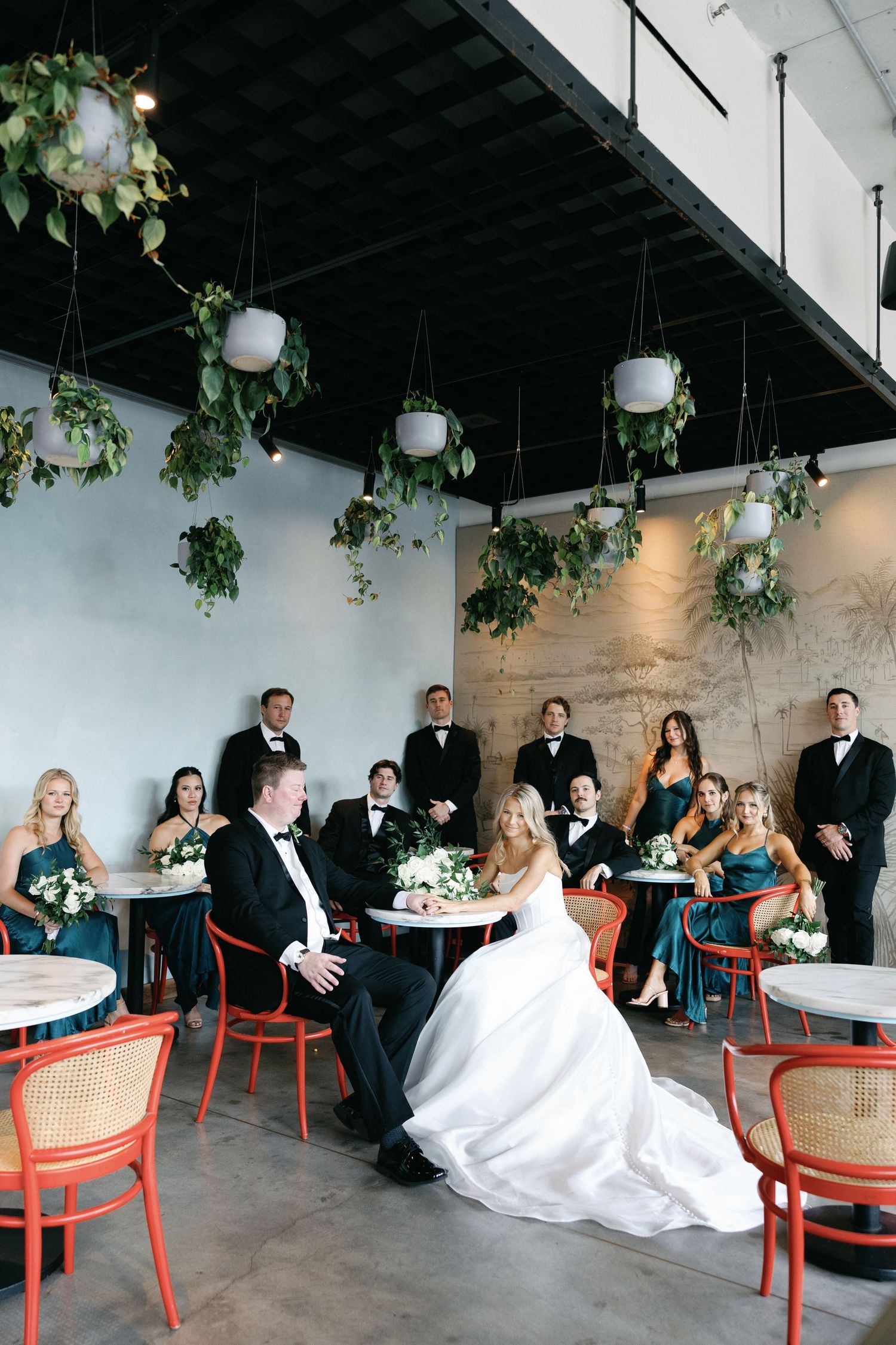 Wedding party relaxes in modern venue with hanging plants as bride and groom sit together at small table.