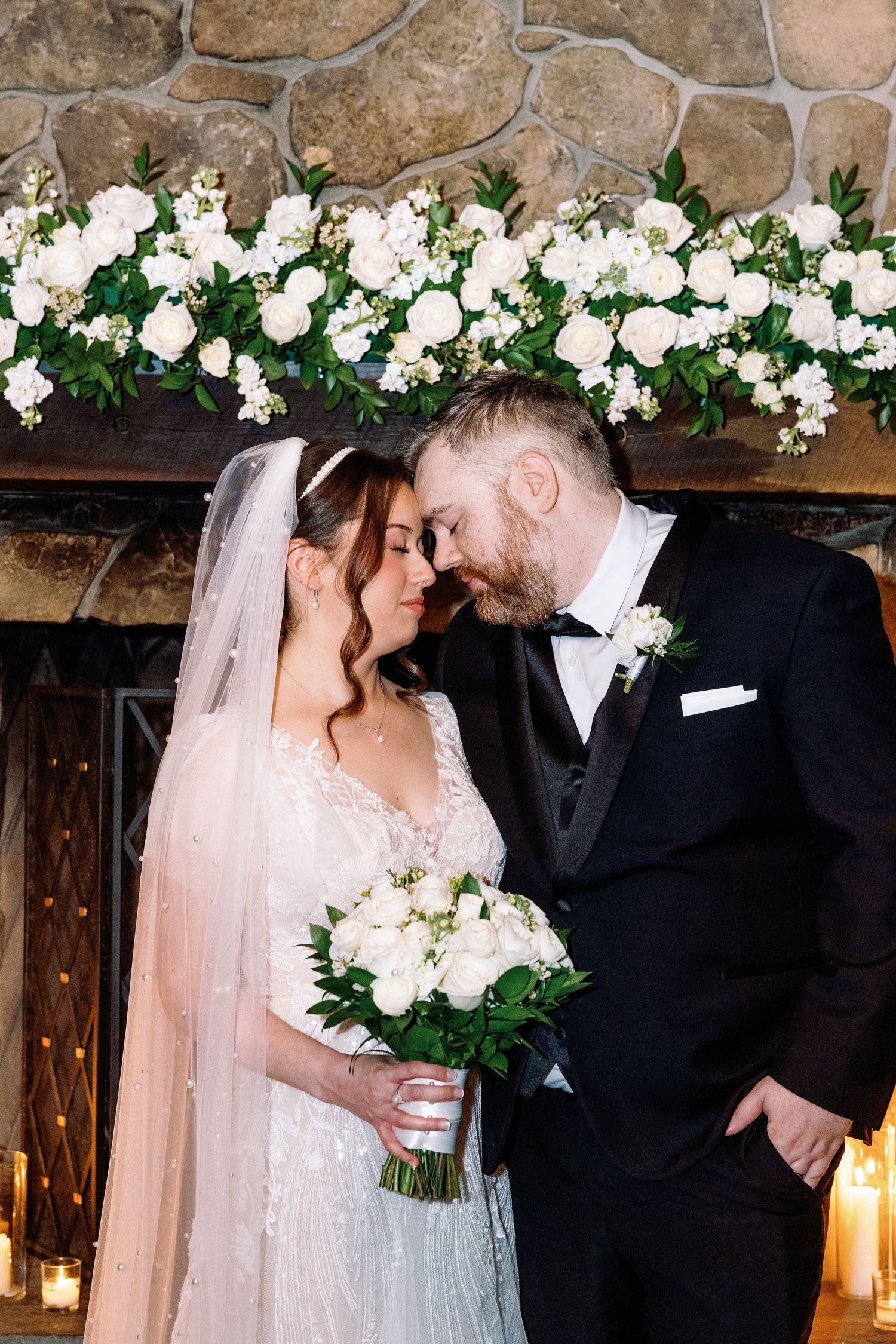 Bride and groom touch foreheads tenderly in front of a stone fireplace adorned with white floral garland.