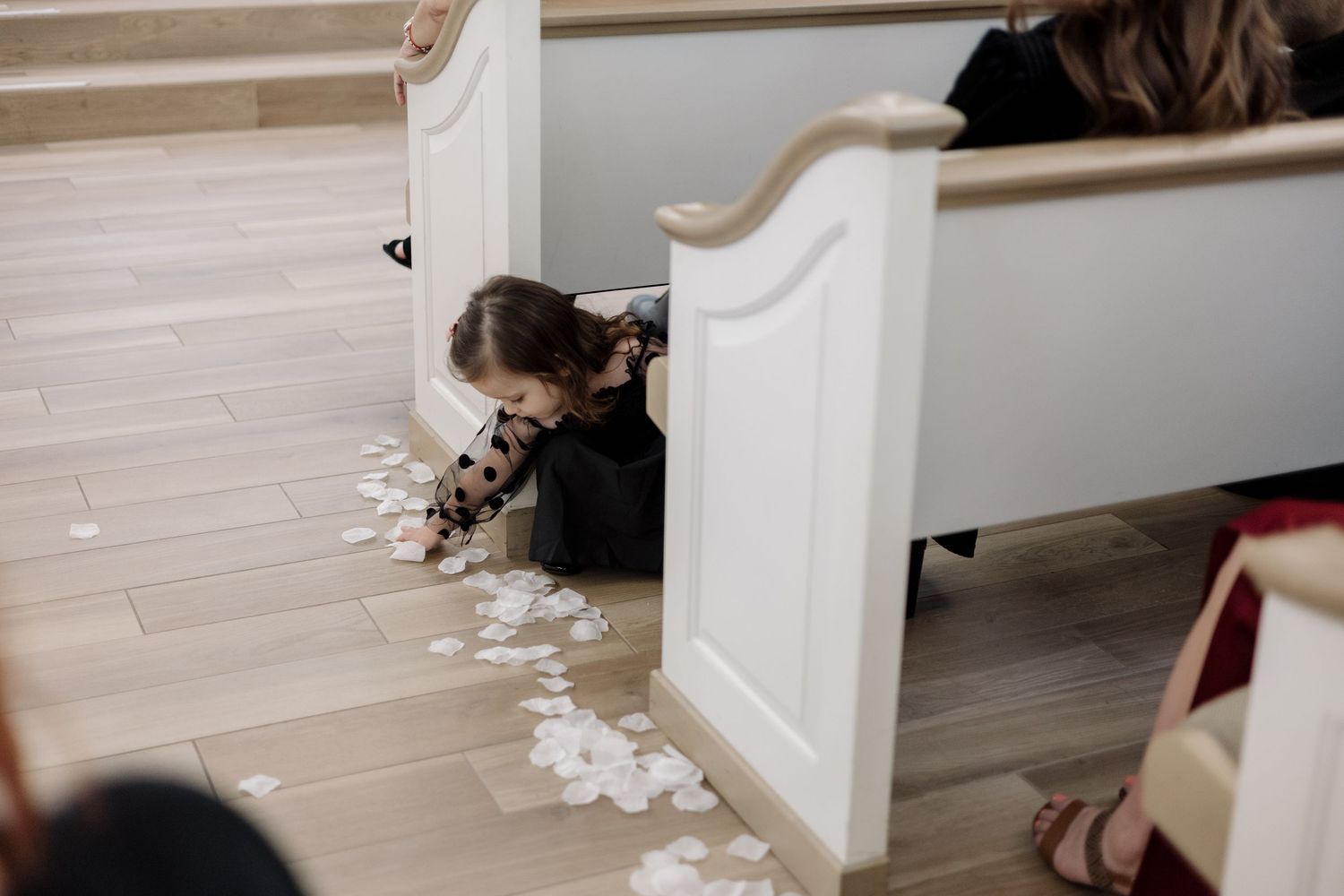 A young child peeks around a white church pew surrounded by scattered flower petals.