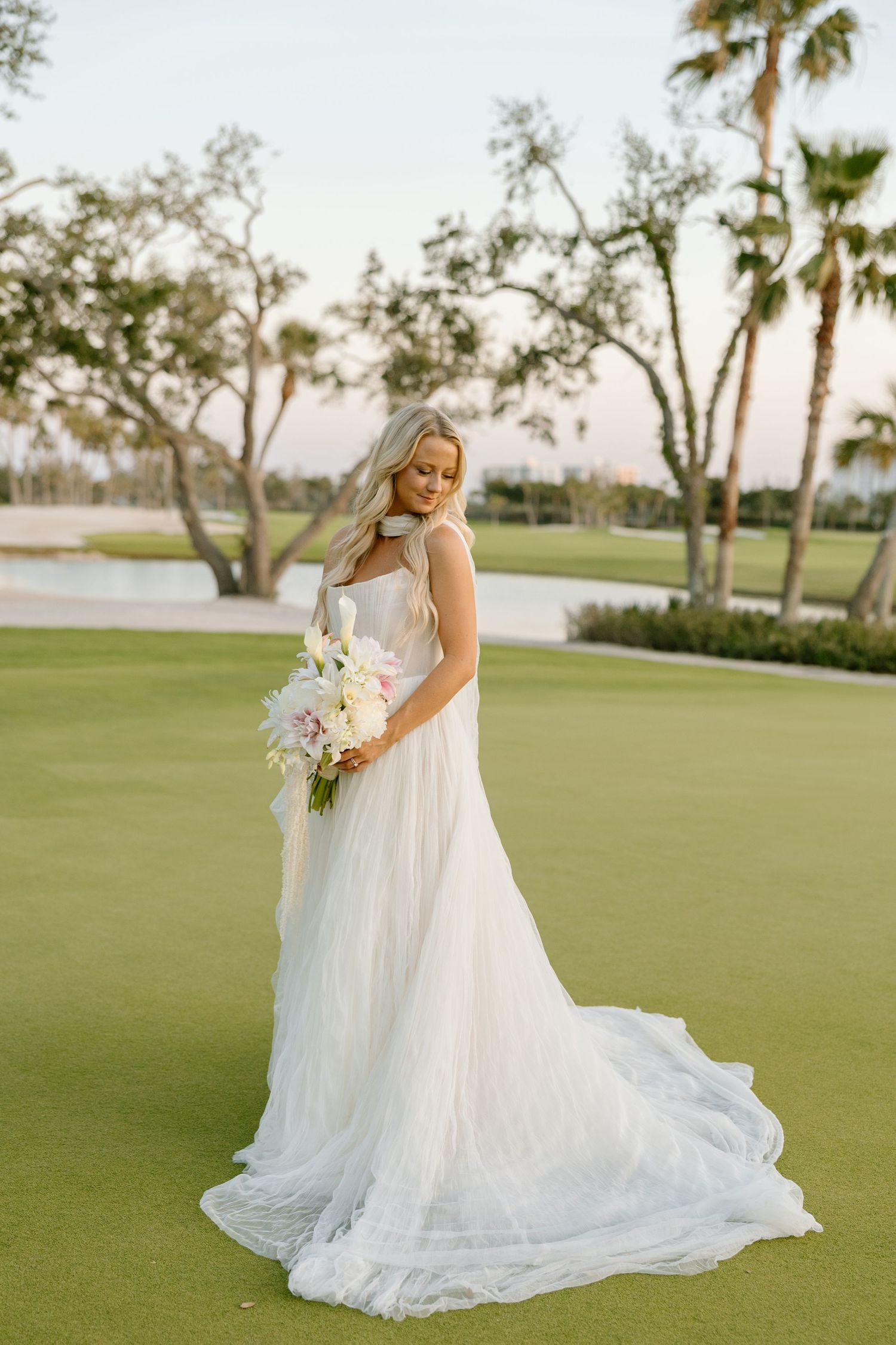 Bride in flowing white tulle gown holding pastel bouquet standing on golf course green at sunset with palm trees.
