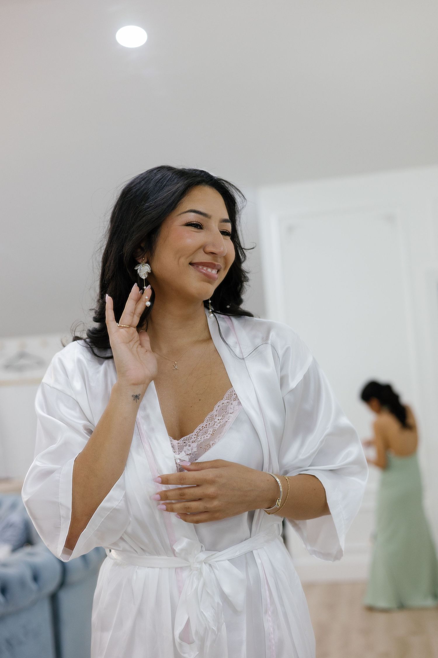Bride in white robe smiles while touching earring during wedding morning preparations.
