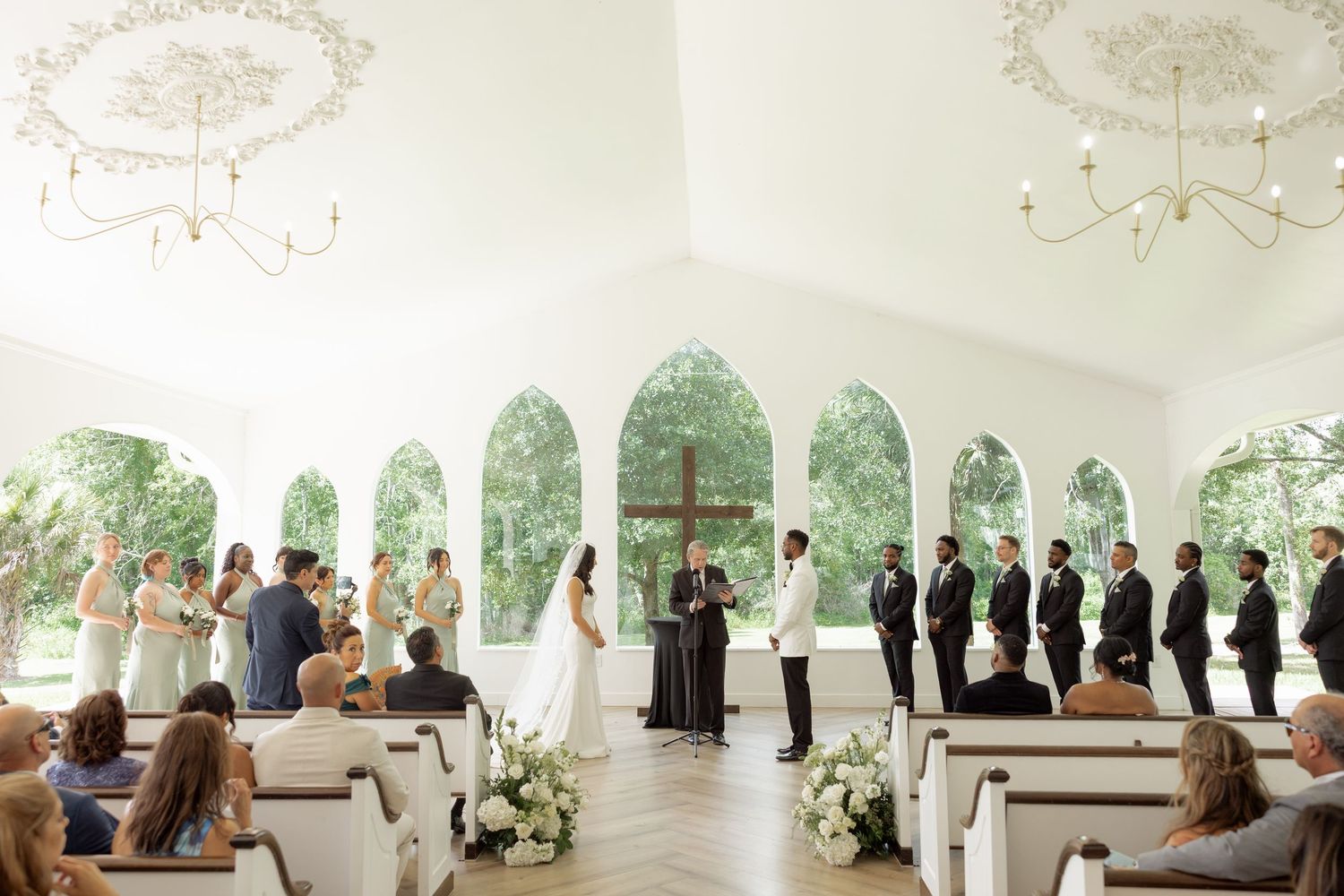 Bride and groom exchange vows at altar inside bright white chapel with arched windows.