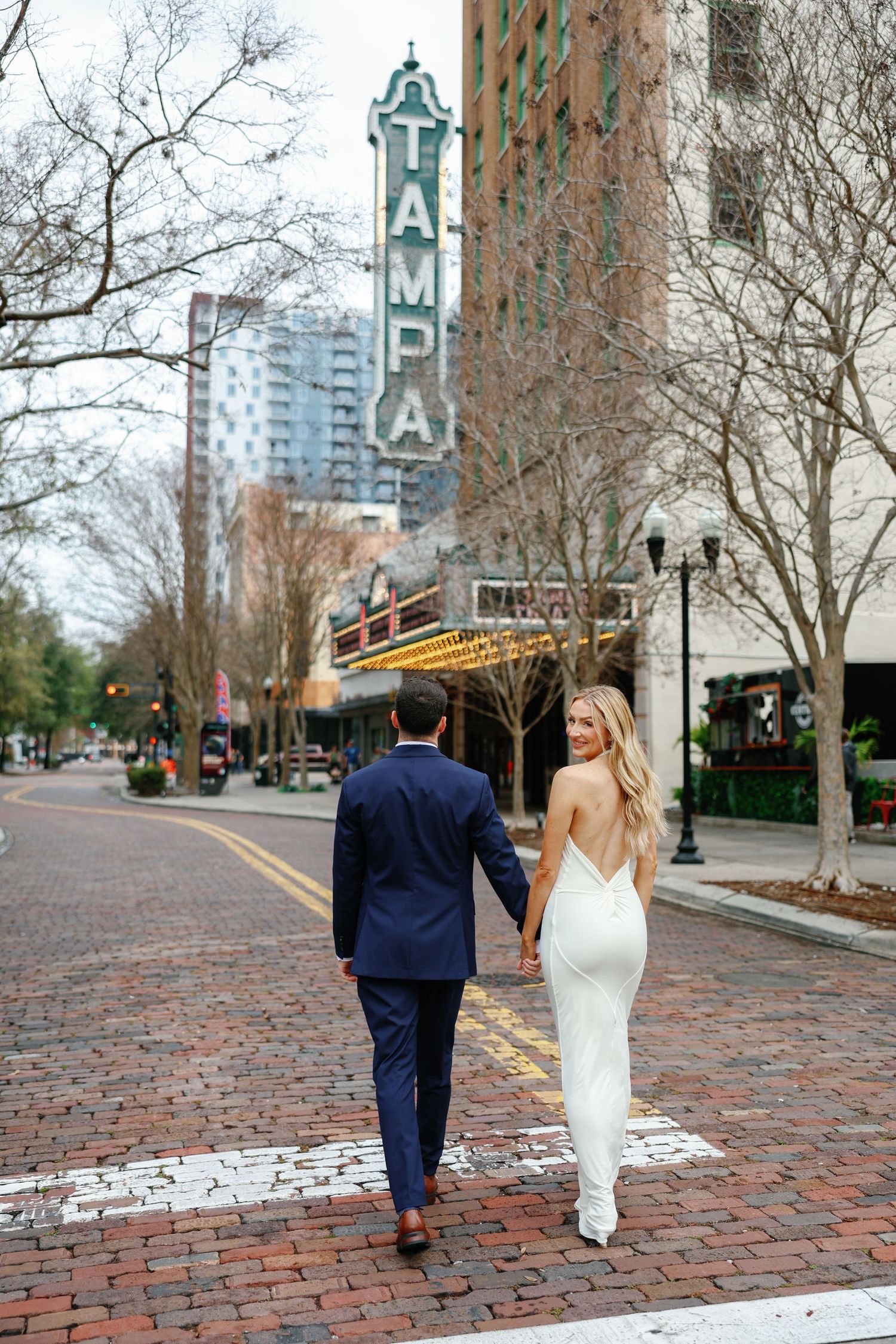 Bride and groom walking hand-in-hand across brick street near iconic Tampa Theatre marquee sign.