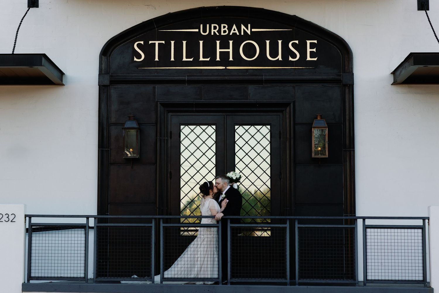 Bride and groom kiss on balcony in front of Urban Stillhouse venue entrance with black double doors.