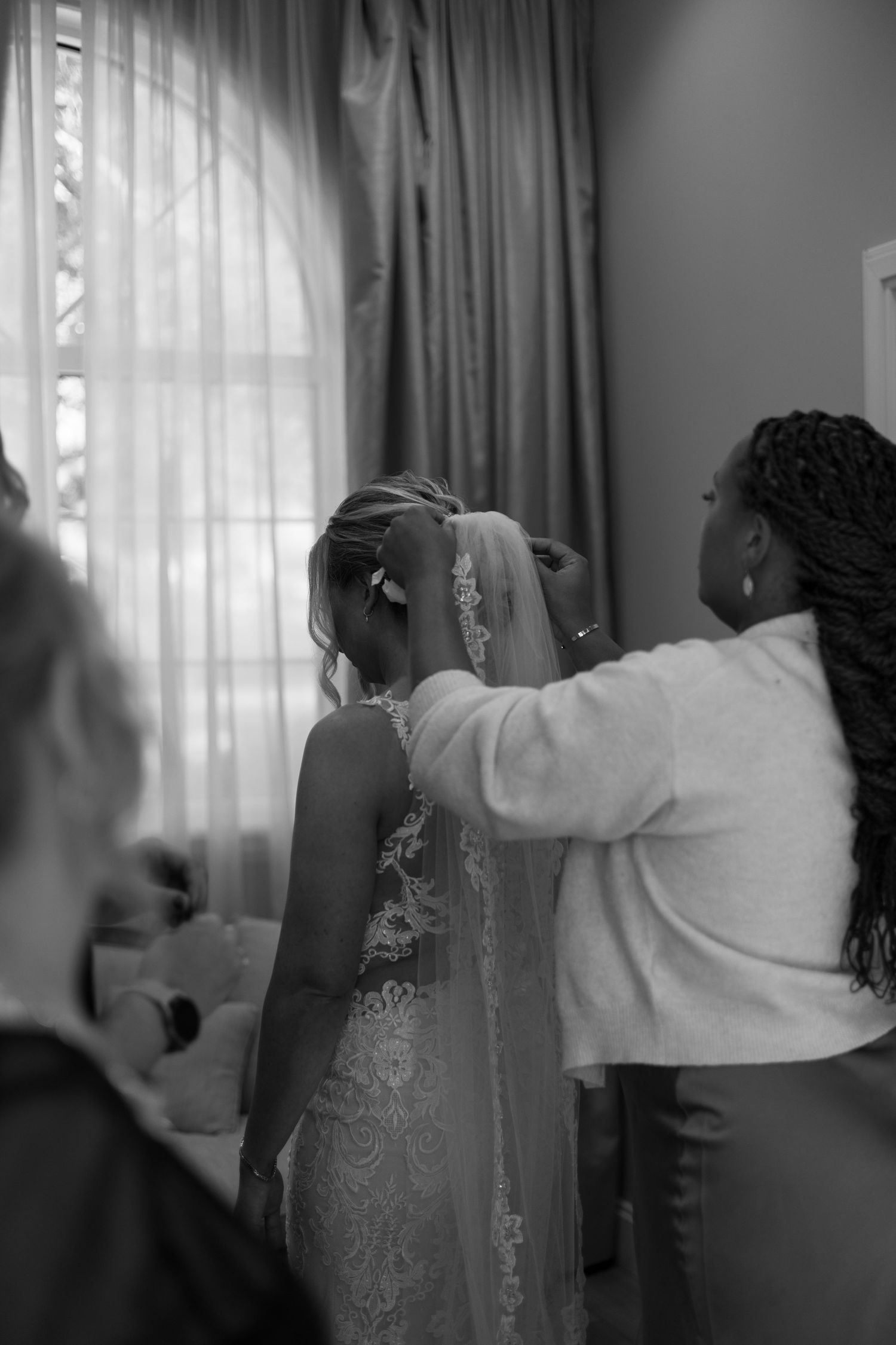Stylist attaches cathedral lace veil to bride's updo hairstyle in softly lit bridal suite.