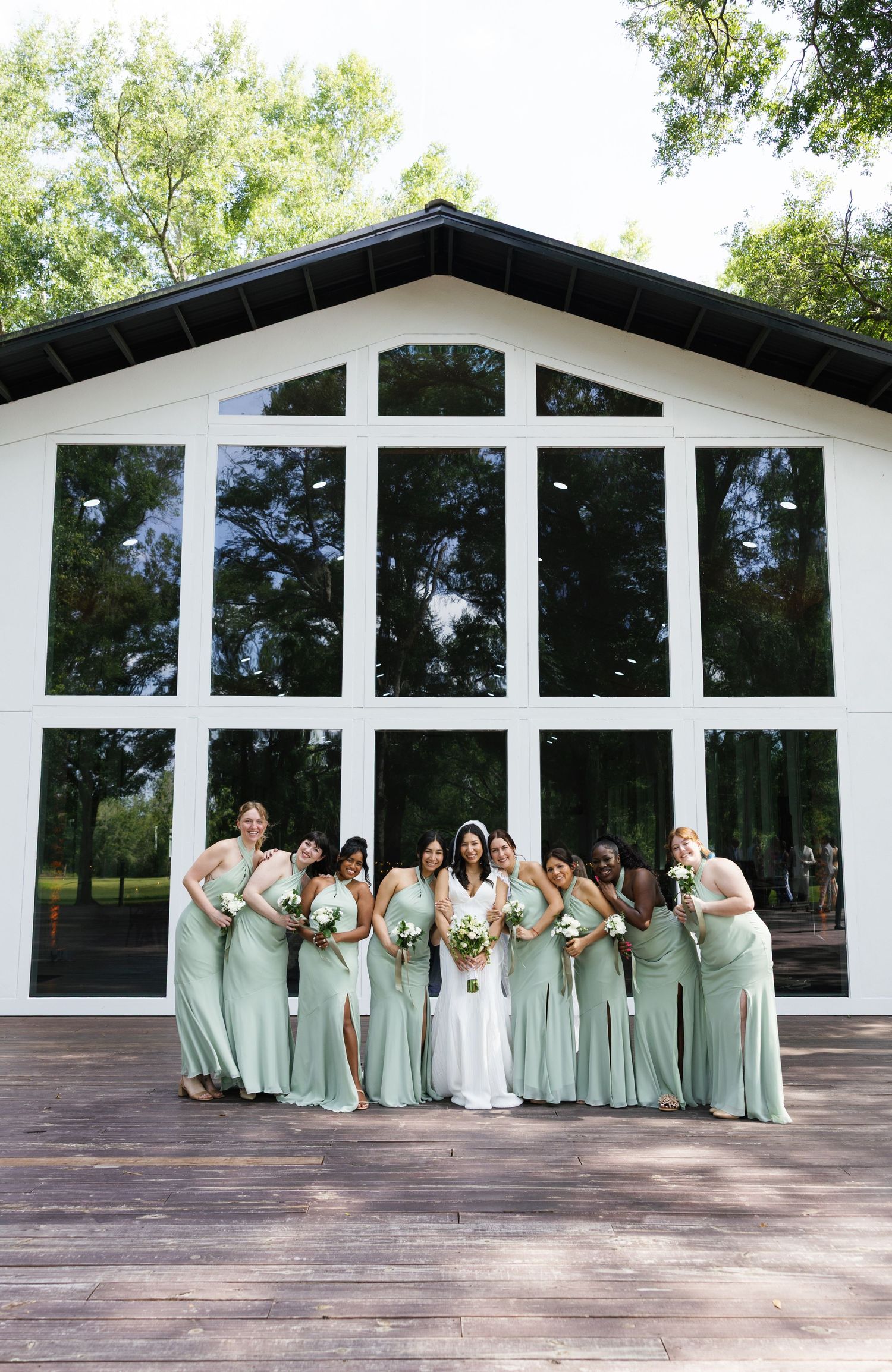 Bride with bridesmaids in sage green gowns pose before large glass wall wedding venue.