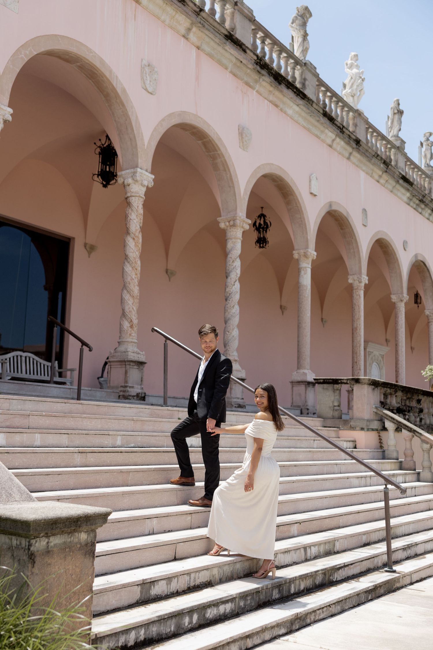 An elegant couple holds hands on marble steps of a grand pink Italian Renaissance-style building outdoors.