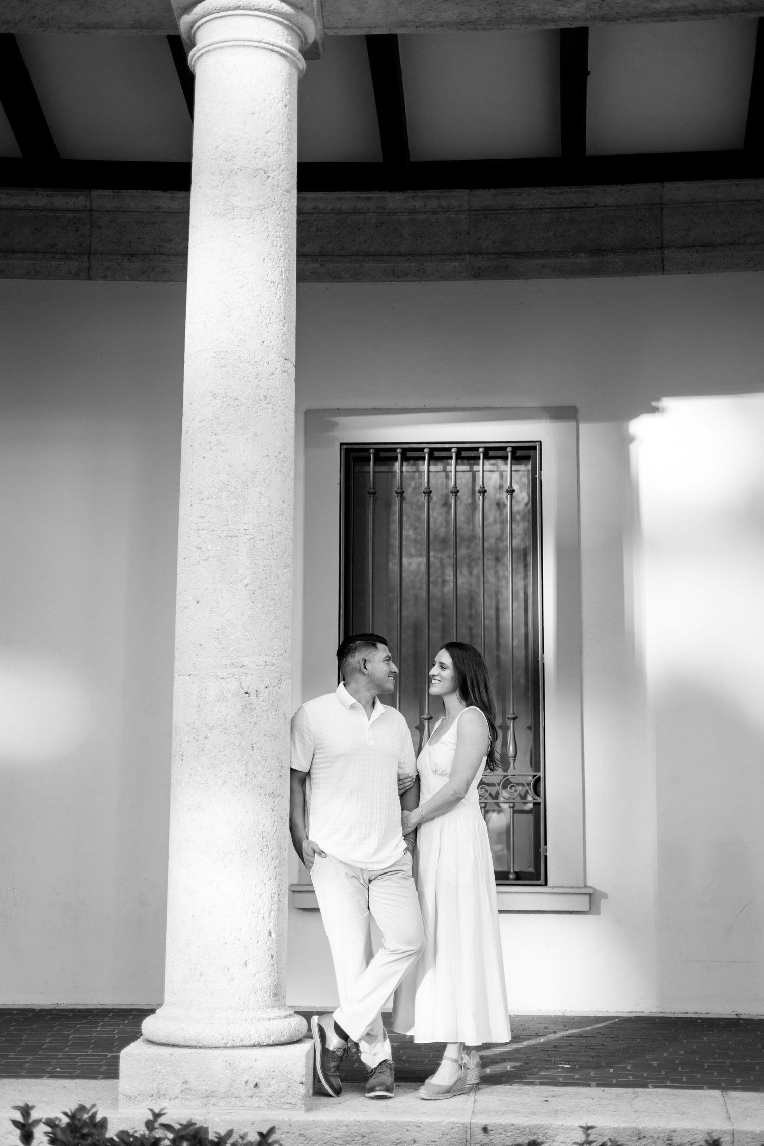 Black and white photo of couple holding hands and gazing at each other beside elegant white column.