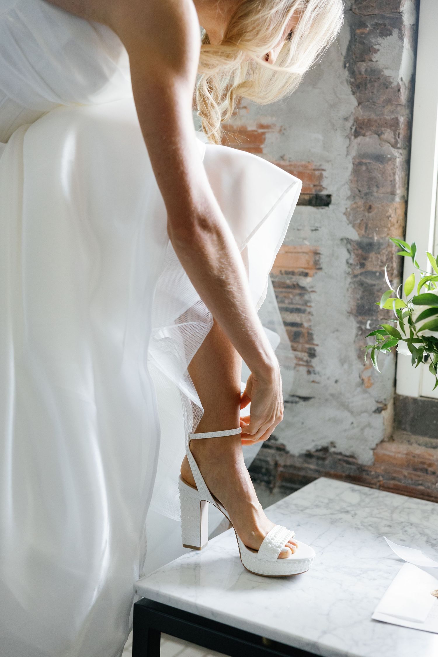 Bride in white strapless gown fastens strappy white block heel shoe while seated near brick wall.
