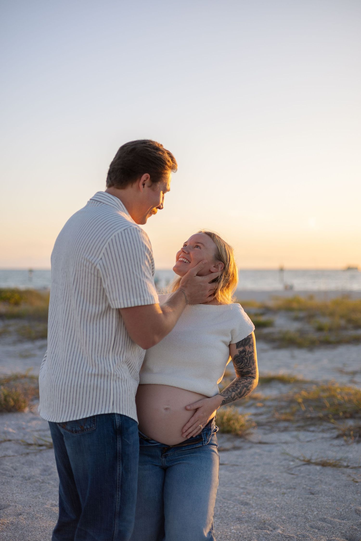 Couple sharing tender moment at sunset beach, man cupping pregnant womans face lovingly.