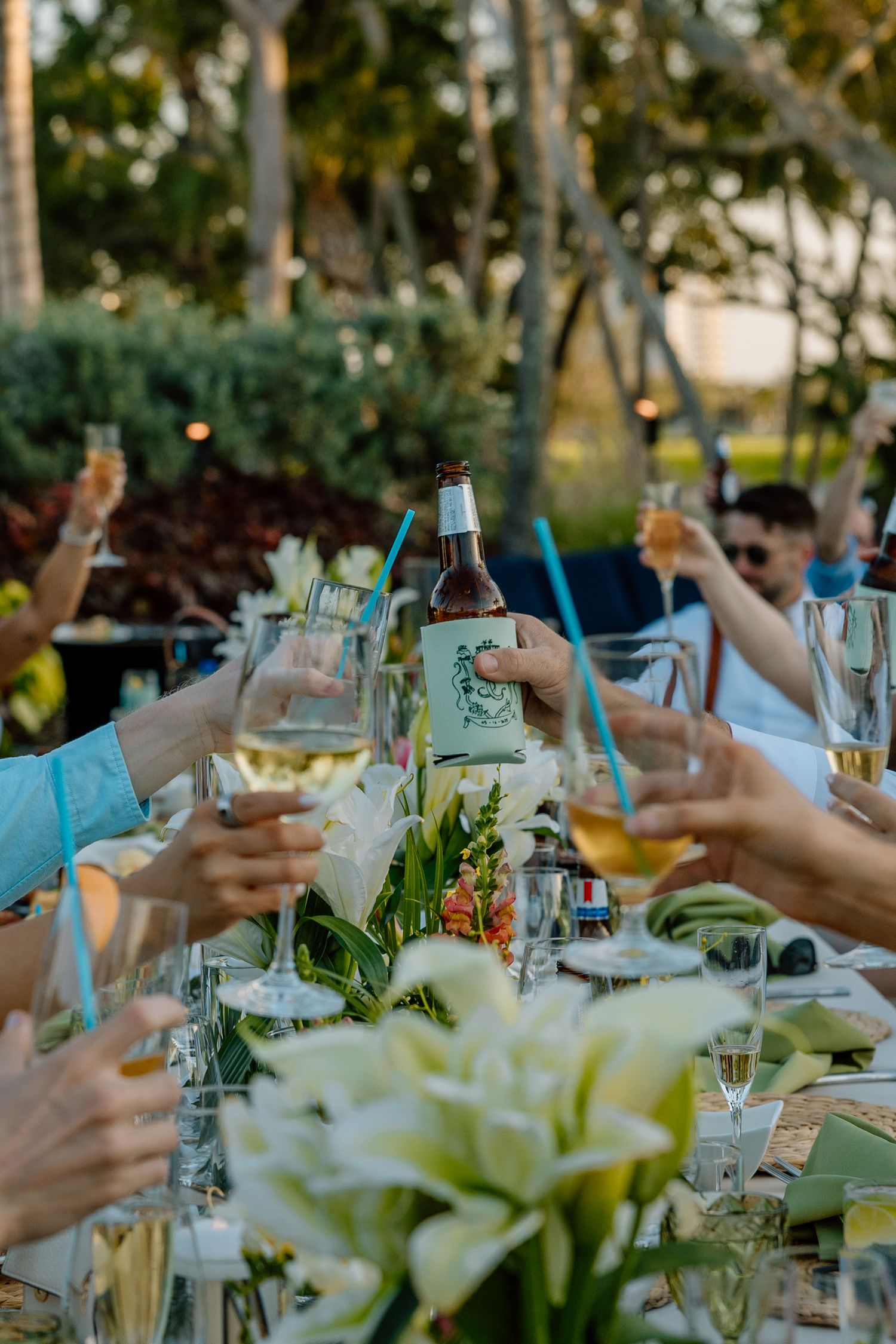 Wedding guests raising glasses and beer bottles in a toast at outdoor reception table adorned with white lilies.