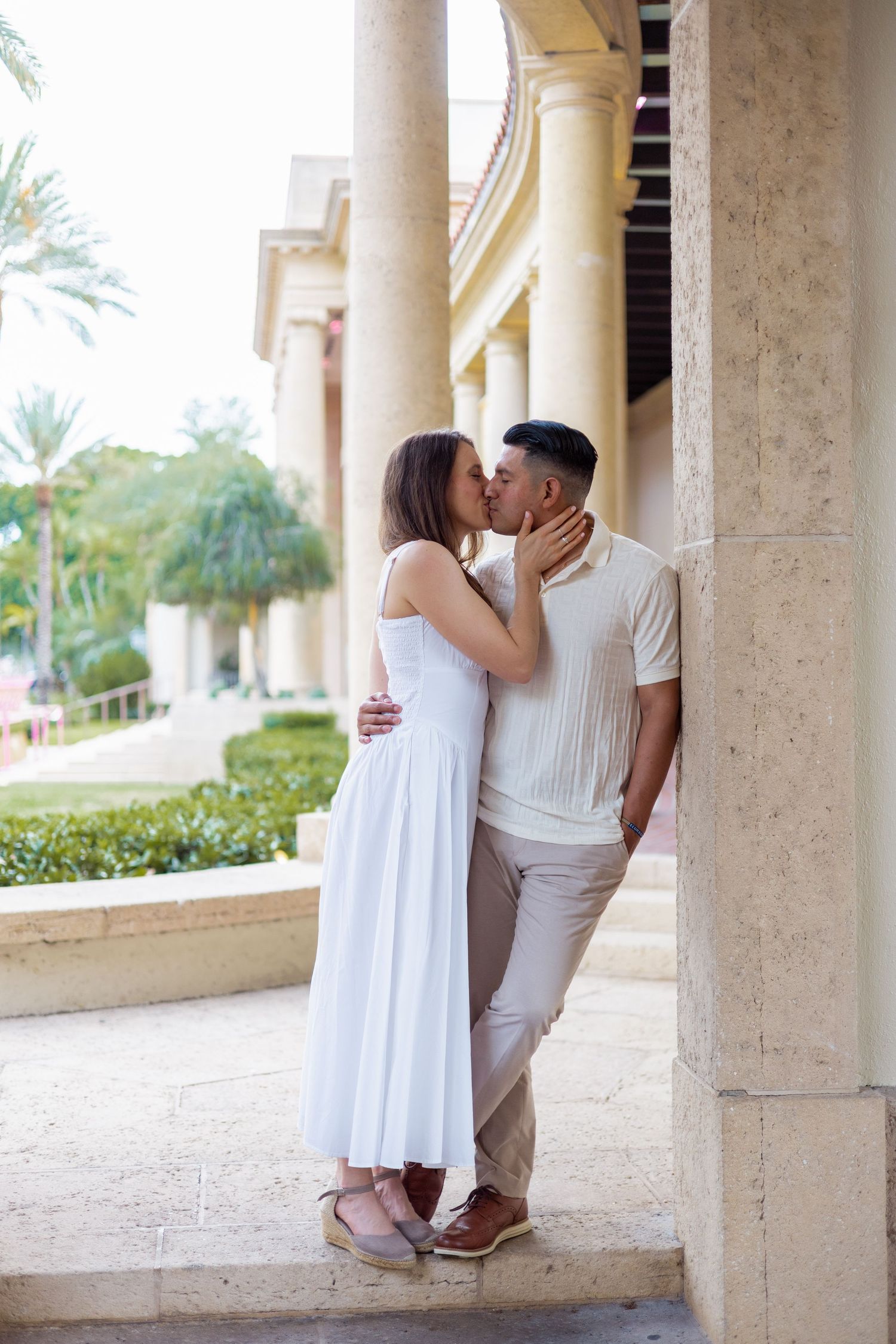 Couple sharing a passionate kiss against a marble column at an elegant outdoor colonnade with palm trees.
