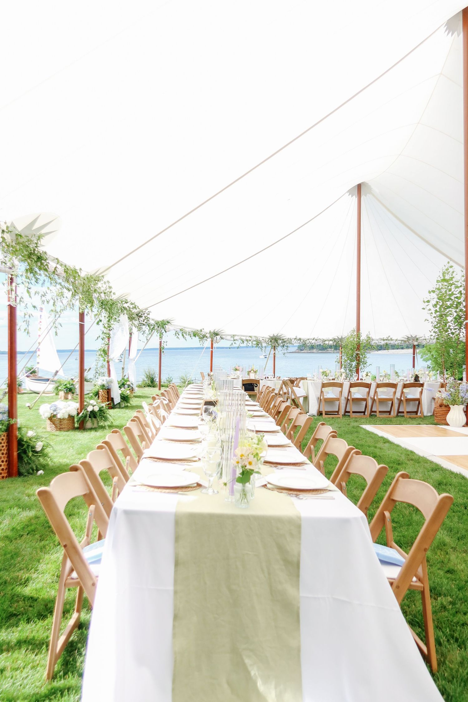 Elegant outdoor wedding reception with long banquet table, wooden chairs, and waterfront view under white sailcloth tent.