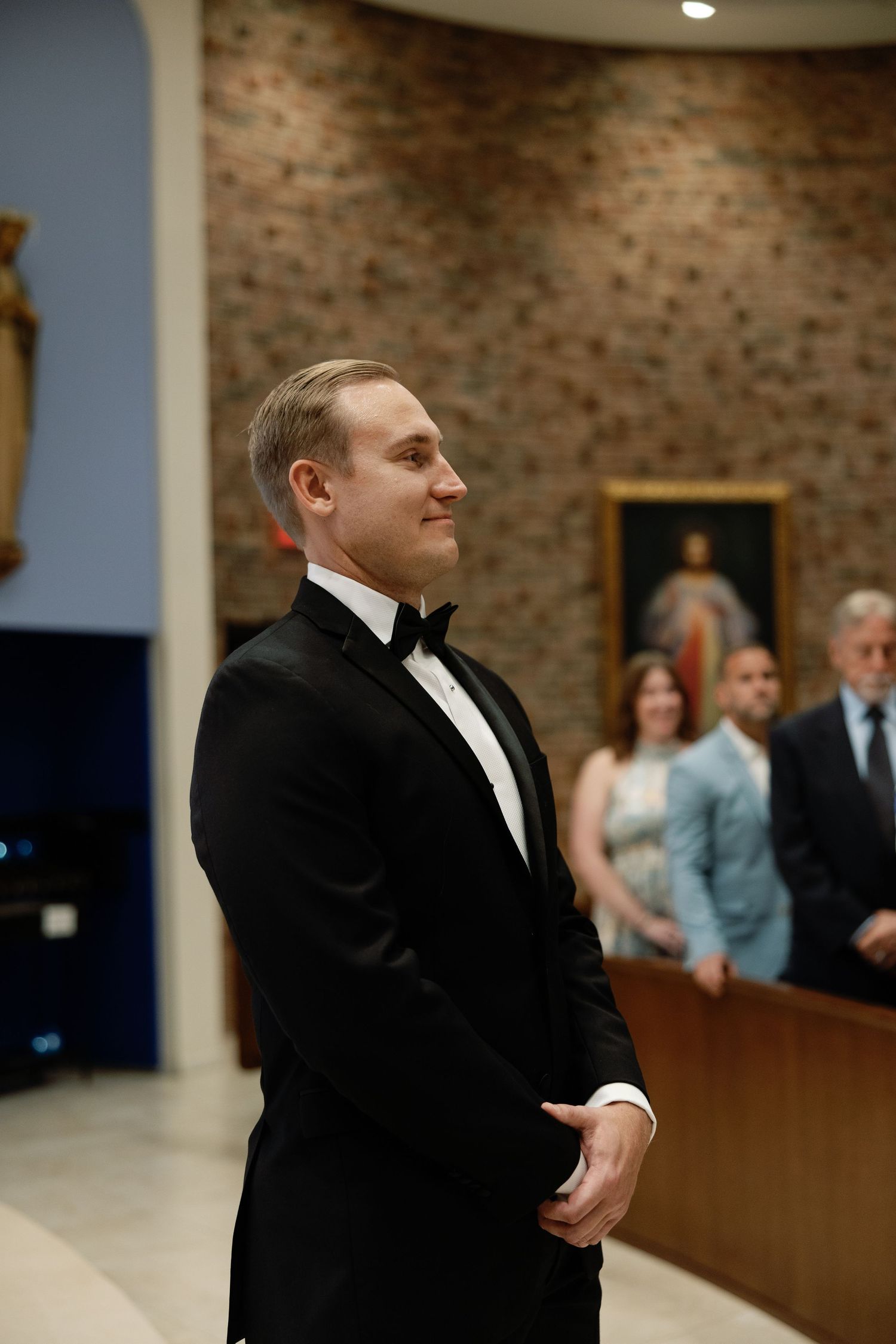 Groom in black tuxedo smiling at altar during wedding ceremony in brick church.