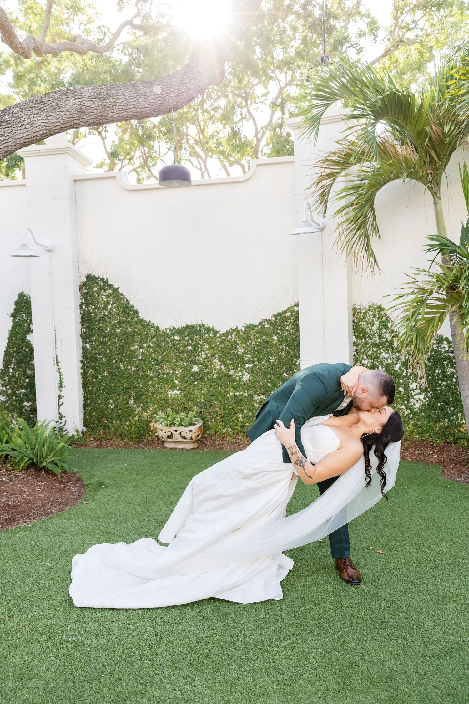 Groom dips bride for a romantic kiss in a lush garden with white walls and tropical greenery at their wedding.