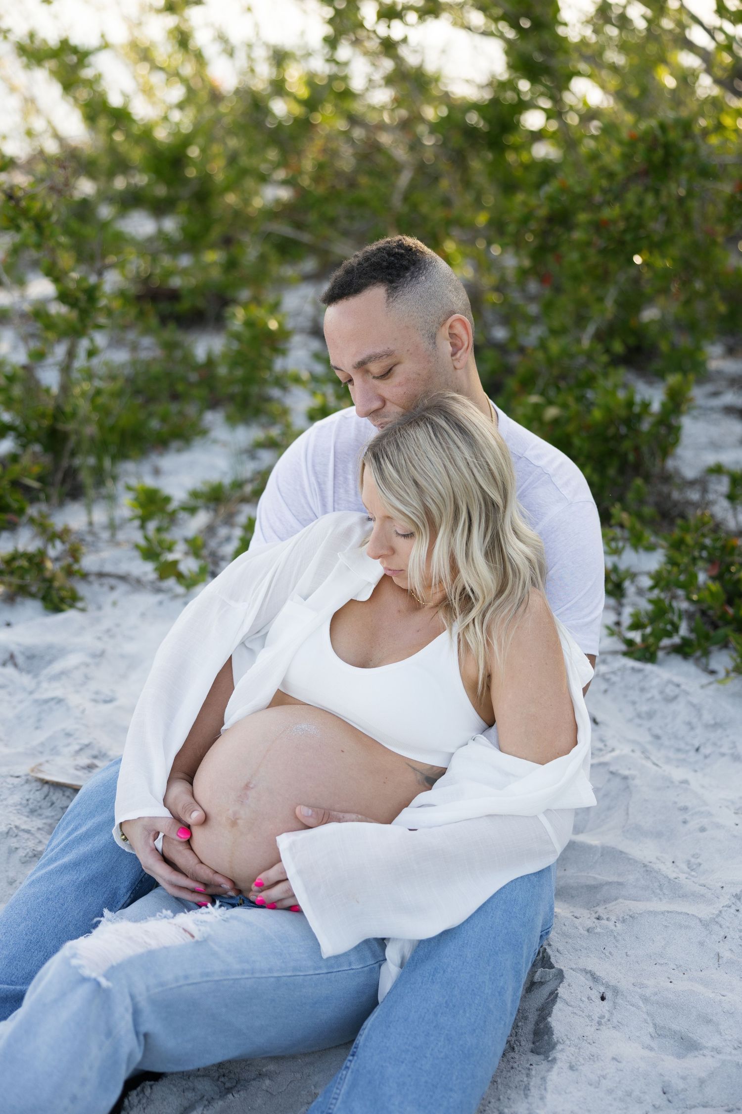 Couple sitting on sandy beach, man embracing pregnant woman from behind at golden hour.
