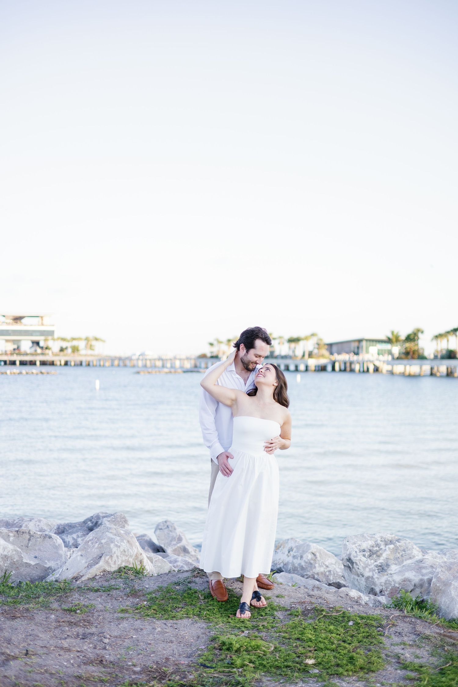 Romantic couple embracing by the waterfront at sunset, woman in white dress leaning back in man's arms.