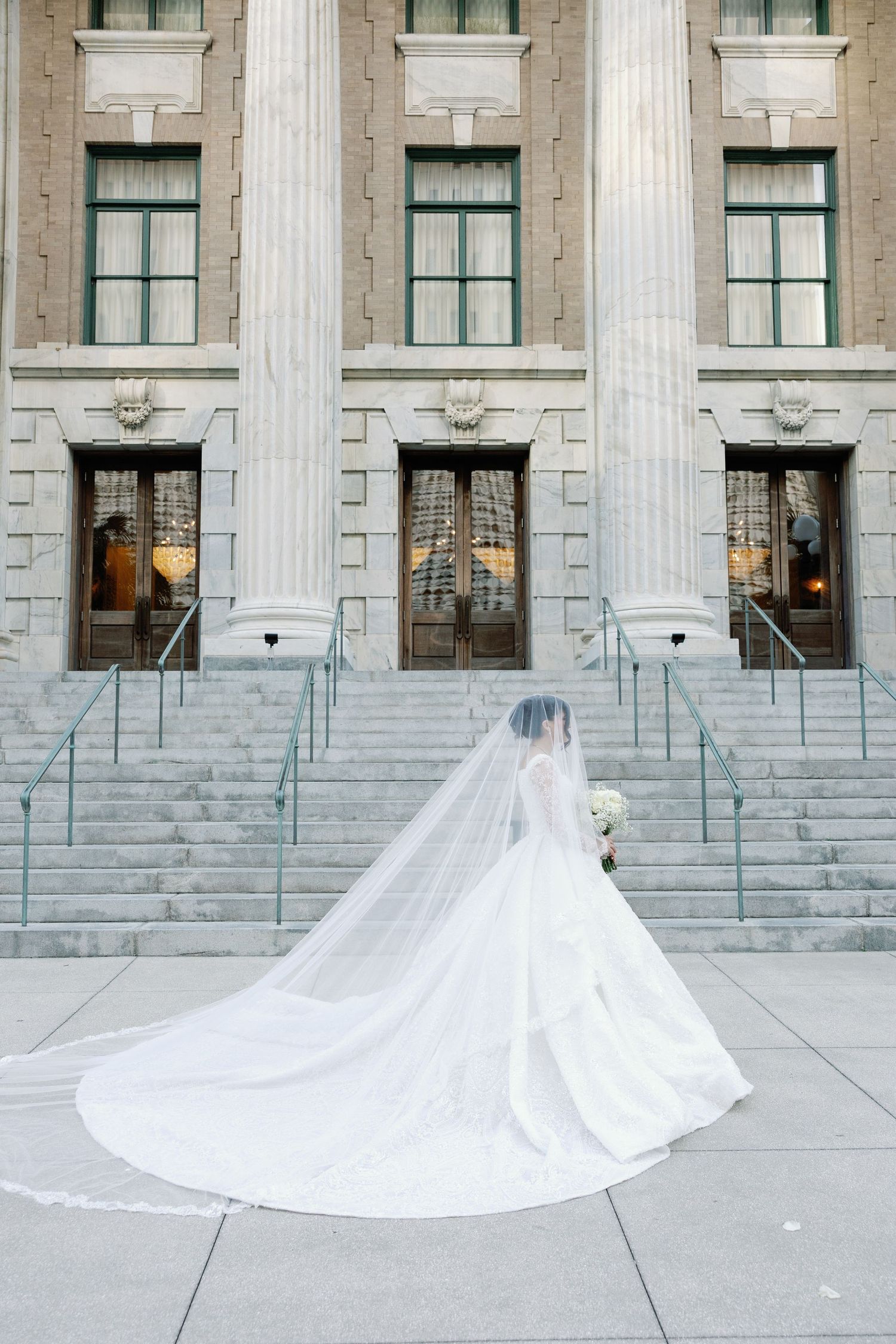 Bride in stunning ballgown with cathedral veil standing before grand stone building steps.