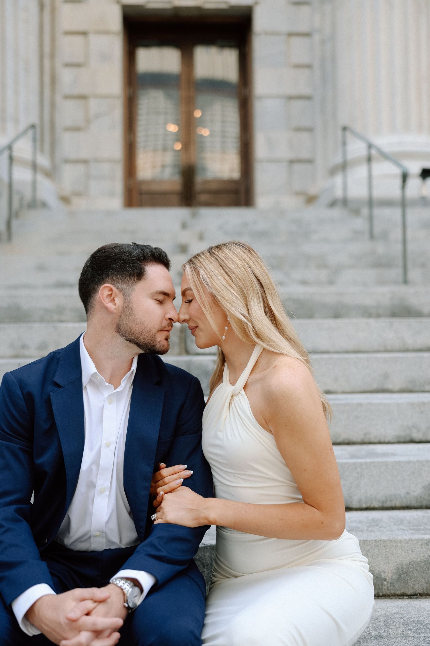 Bride and groom sharing a tender almost-kiss while seated on stone steps of historic building.
