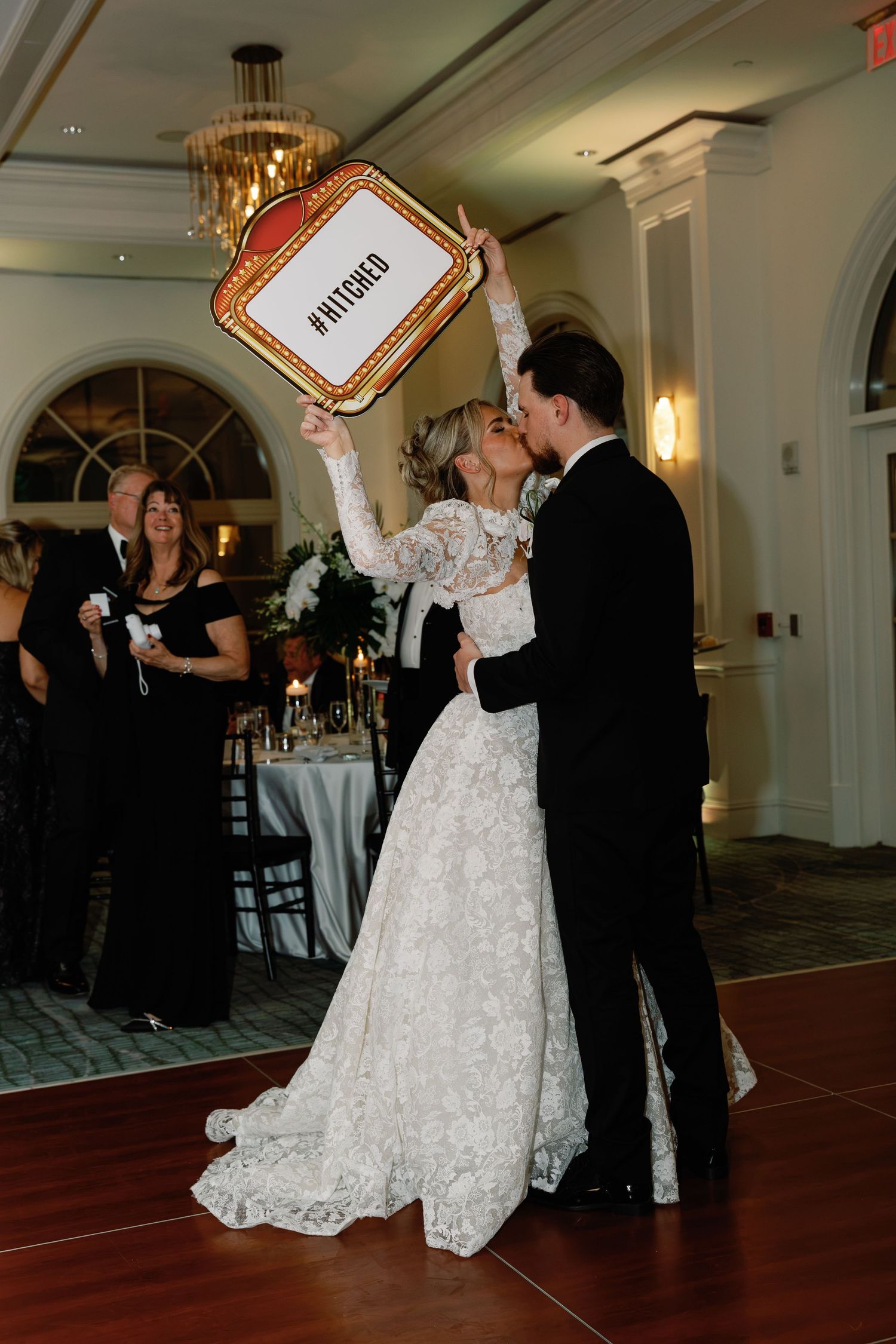 Bride and groom share a kiss on the dance floor, holding a #HITCHED sign at their elegant wedding reception.