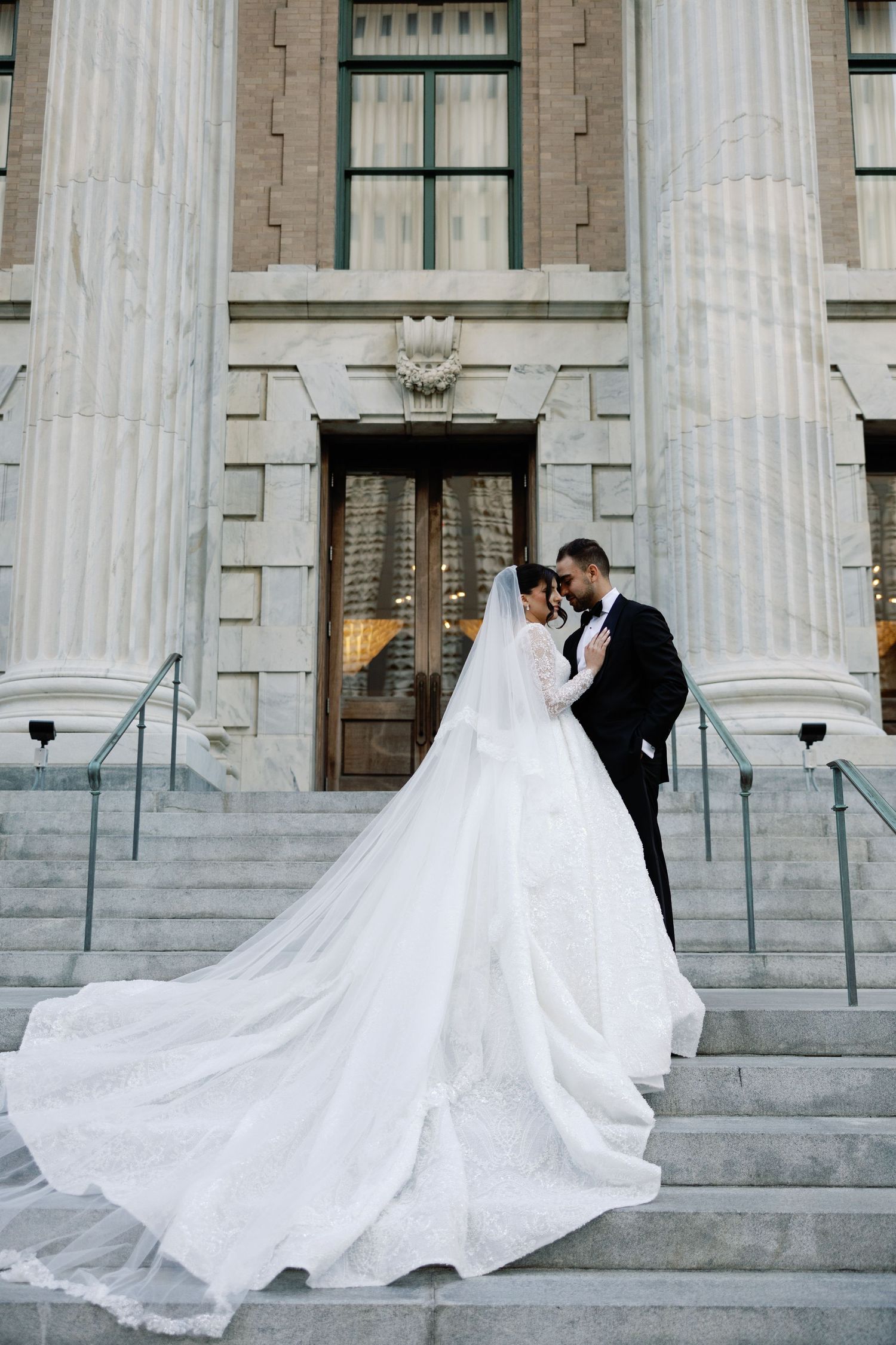 Bride in elegant ballgown with long veil and groom in tuxedo embracing on grand stone steps.