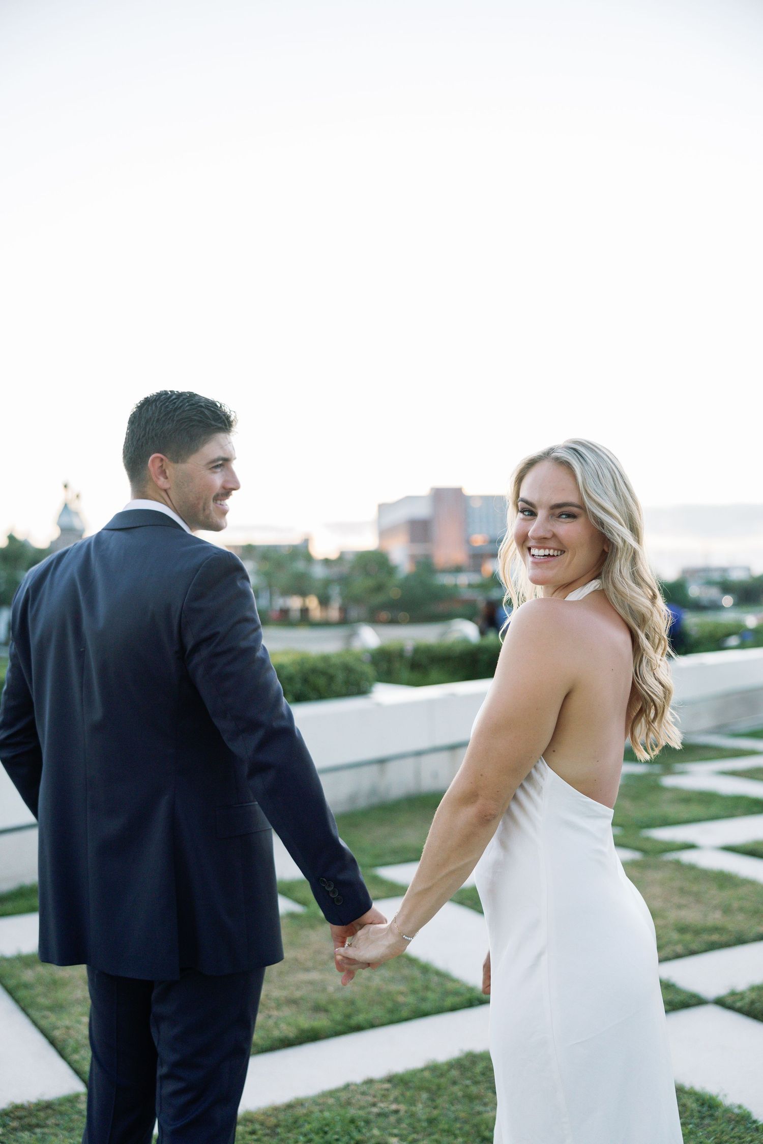 Smiling blonde bride in halter gown looks back while holding hands with groom on rooftop terrace.