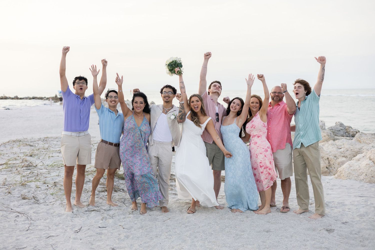 Happy wedding party celebrating on sandy beach, group of ten raising arms in joy with ocean in background.