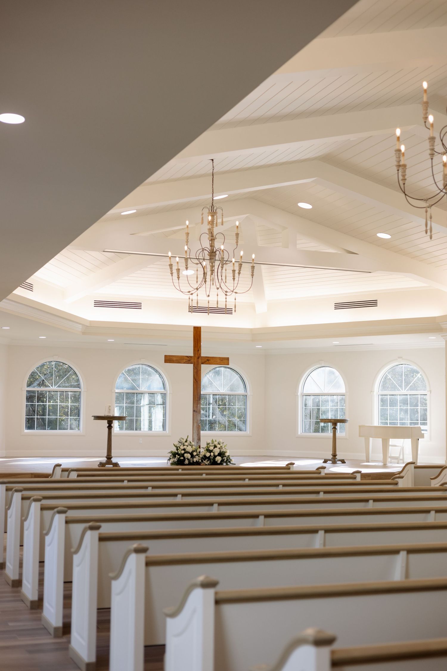 Bright white chapel interior with wooden cross, floral altar arrangement, chandeliers and arched windows.