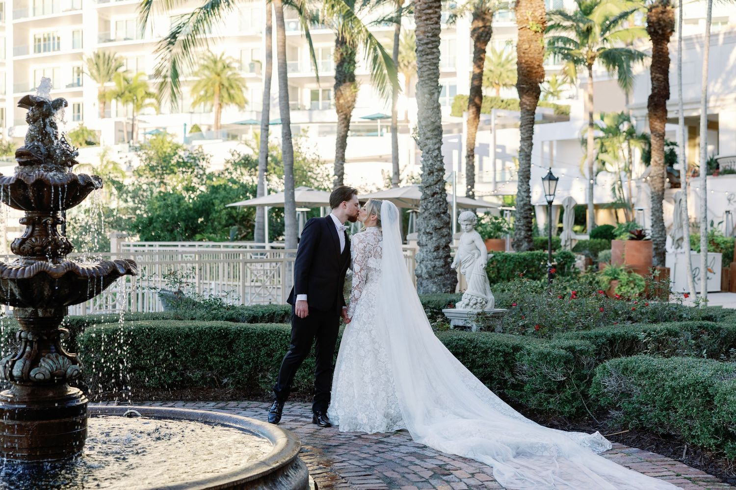 Bride and groom share a romantic kiss near a fountain in a lush garden with palm trees at an elegant venue.