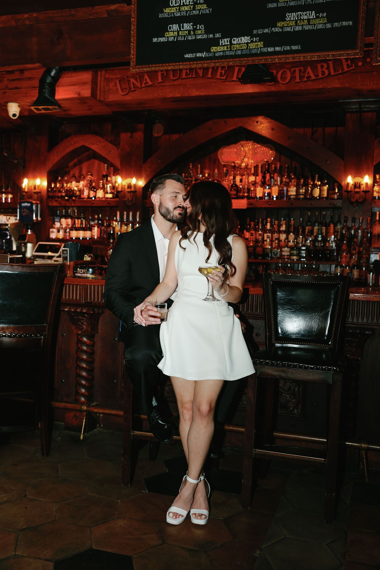 Stylish couple shares a romantic kiss at a warmly lit vintage bar, woman in white mini dress.