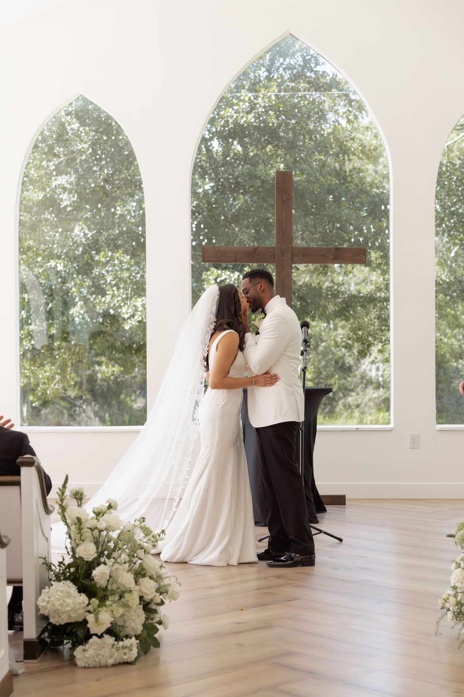 Bride and groom share first kiss at chapel altar with wooden cross and arched windows.