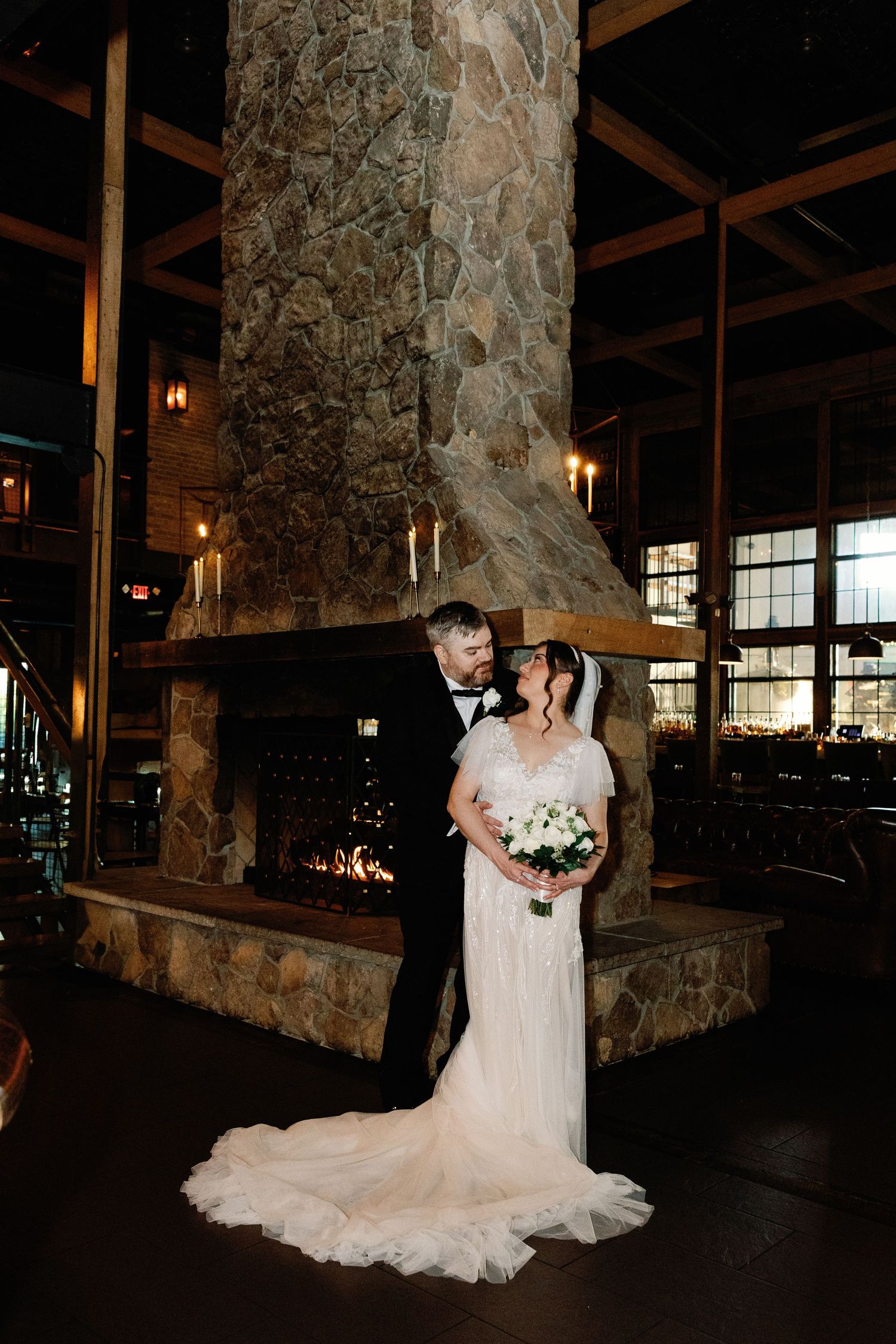 Bride and groom stand before a grand stone fireplace inside a rustic lodge-style wedding reception venue.