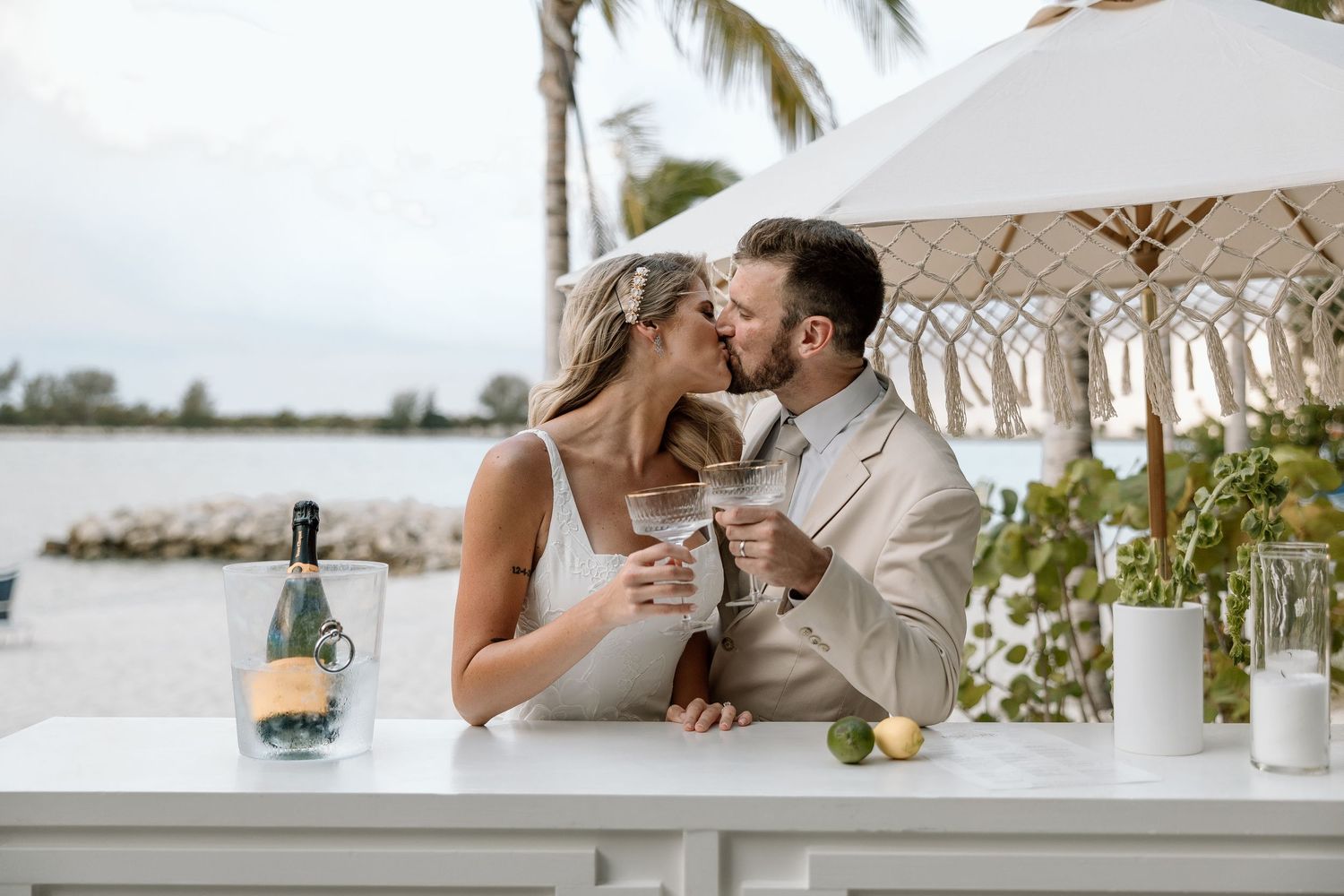 Bride and groom sharing kiss while toasting champagne glasses at outdoor beach bar with tropical waterfront view.
