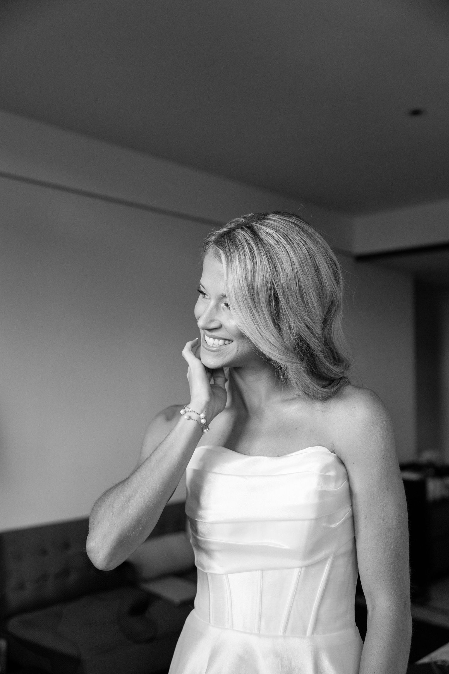 Smiling bride in strapless white wedding gown touching her face in black and white portrait photo.