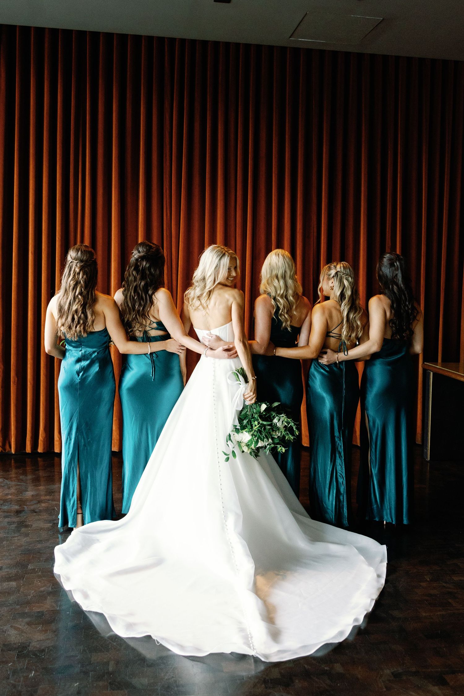 Bride and five bridesmaids in teal satin gowns pose with backs to camera in front of orange curtain.