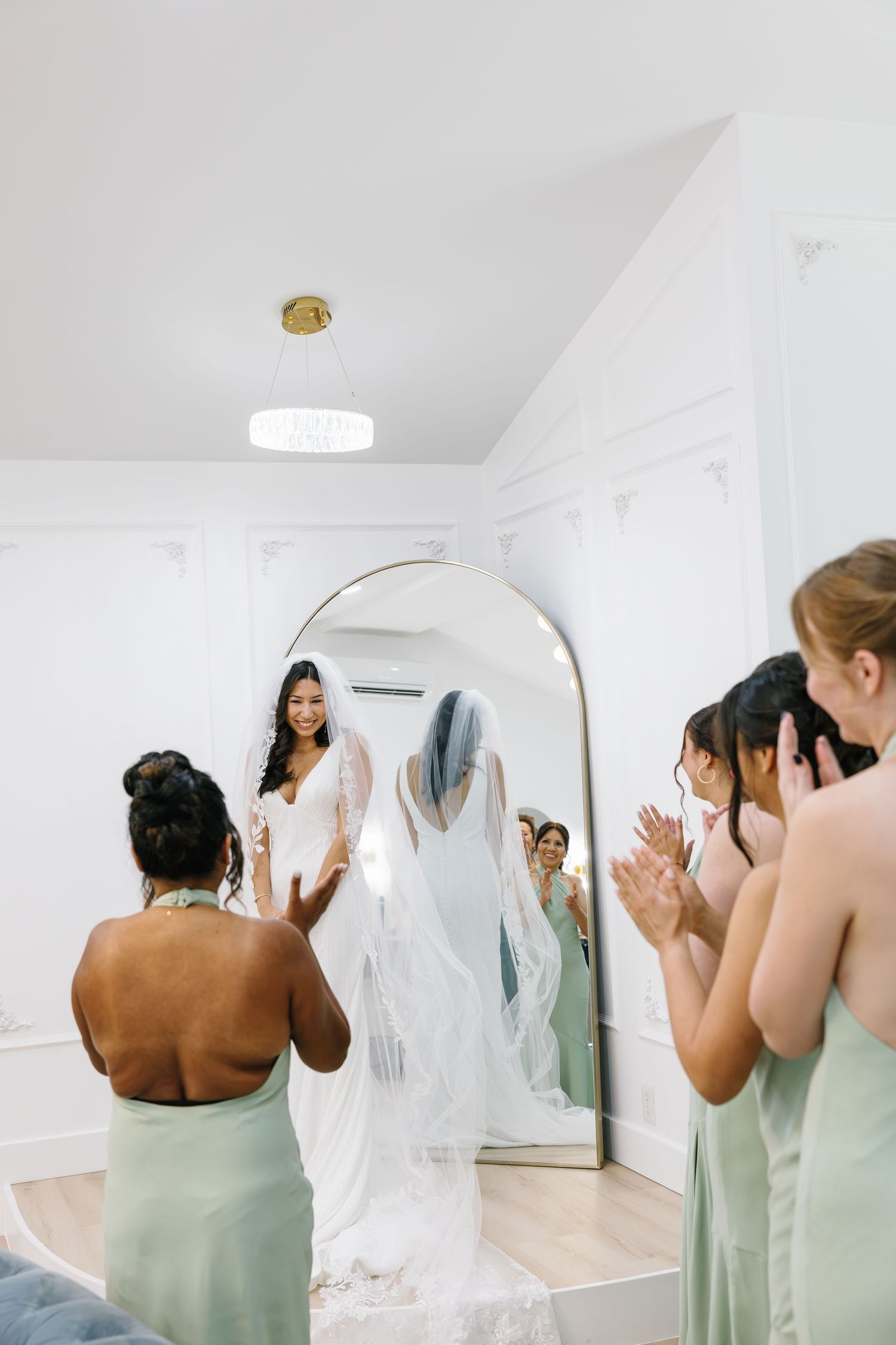 Bride in white gown and blue veil smiles at bridesmaids in sage green dresses clapping in bright white bridal suite.