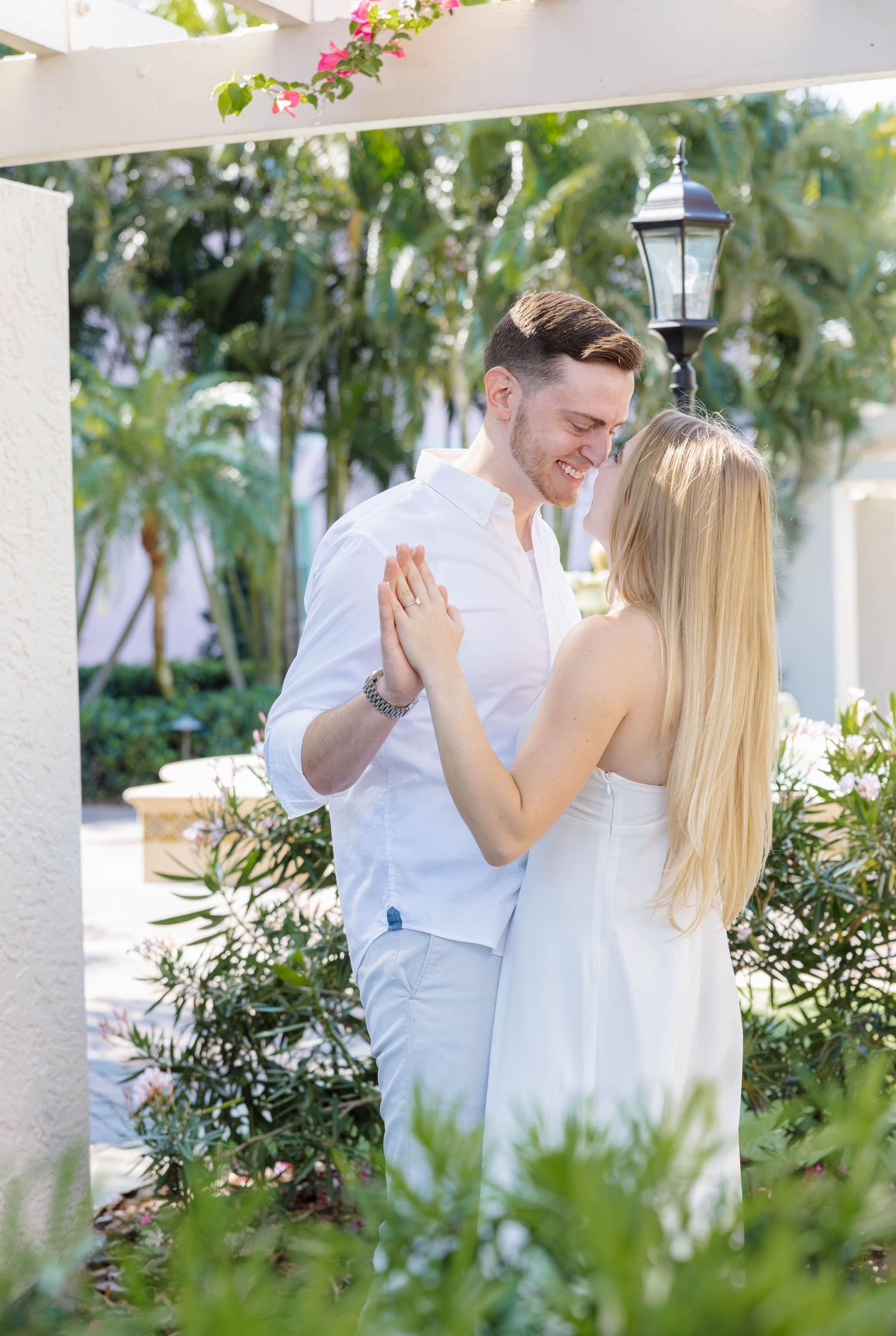 Smiling couple holding hands and facing each other under a sunny garden pergola.