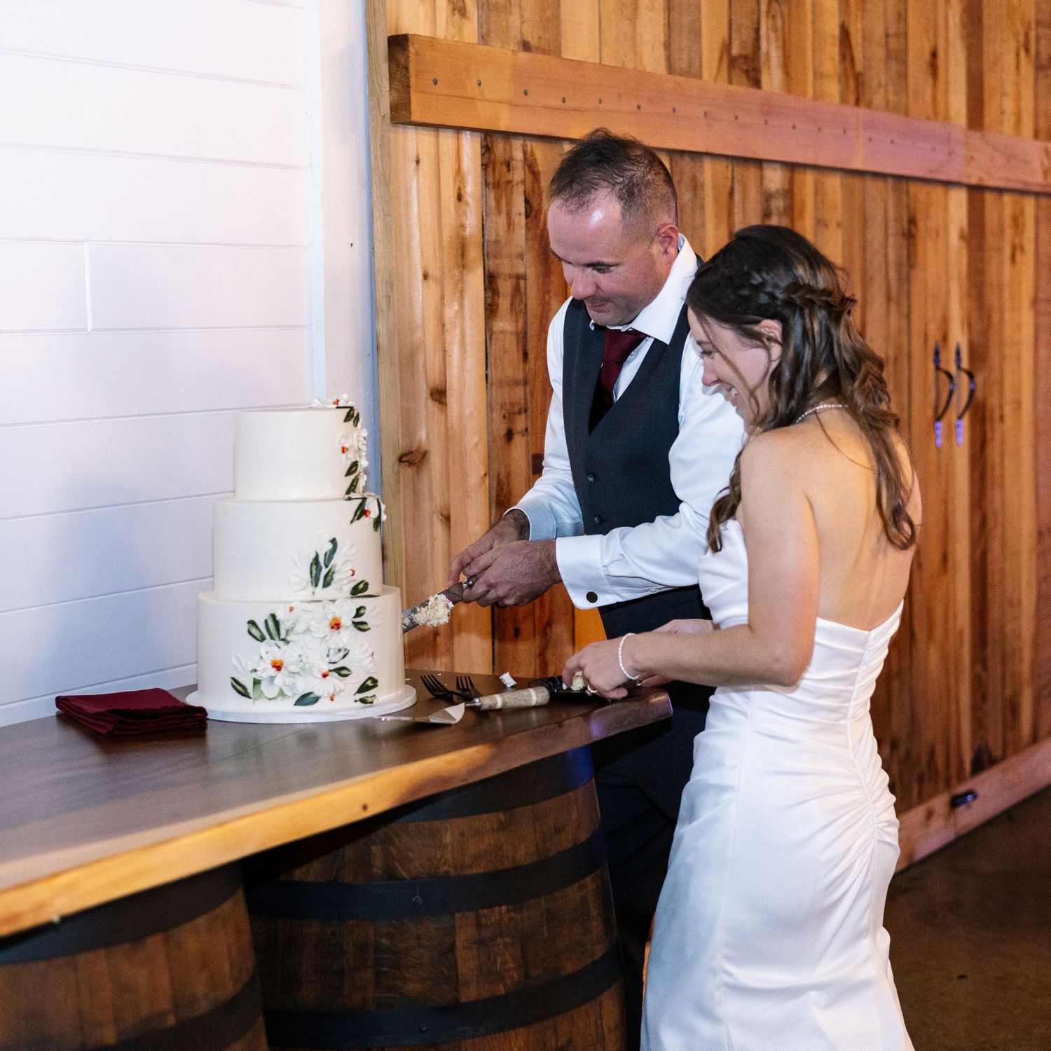 Bride and groom smiling while cutting a elegant white floral wedding cake in a rustic barn venue.
