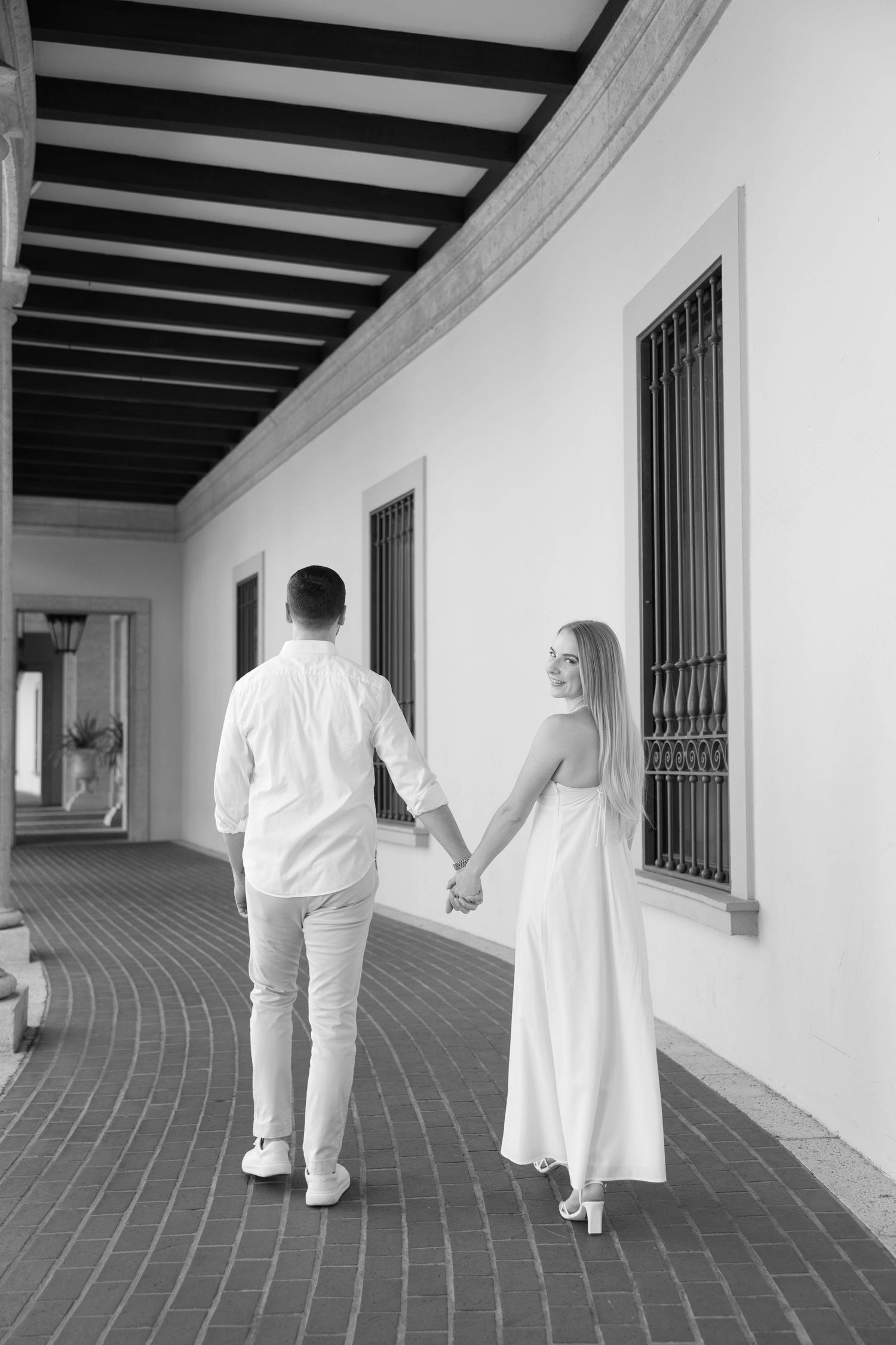 Black and white photo of couple walking hand in hand through an elegant corridor.